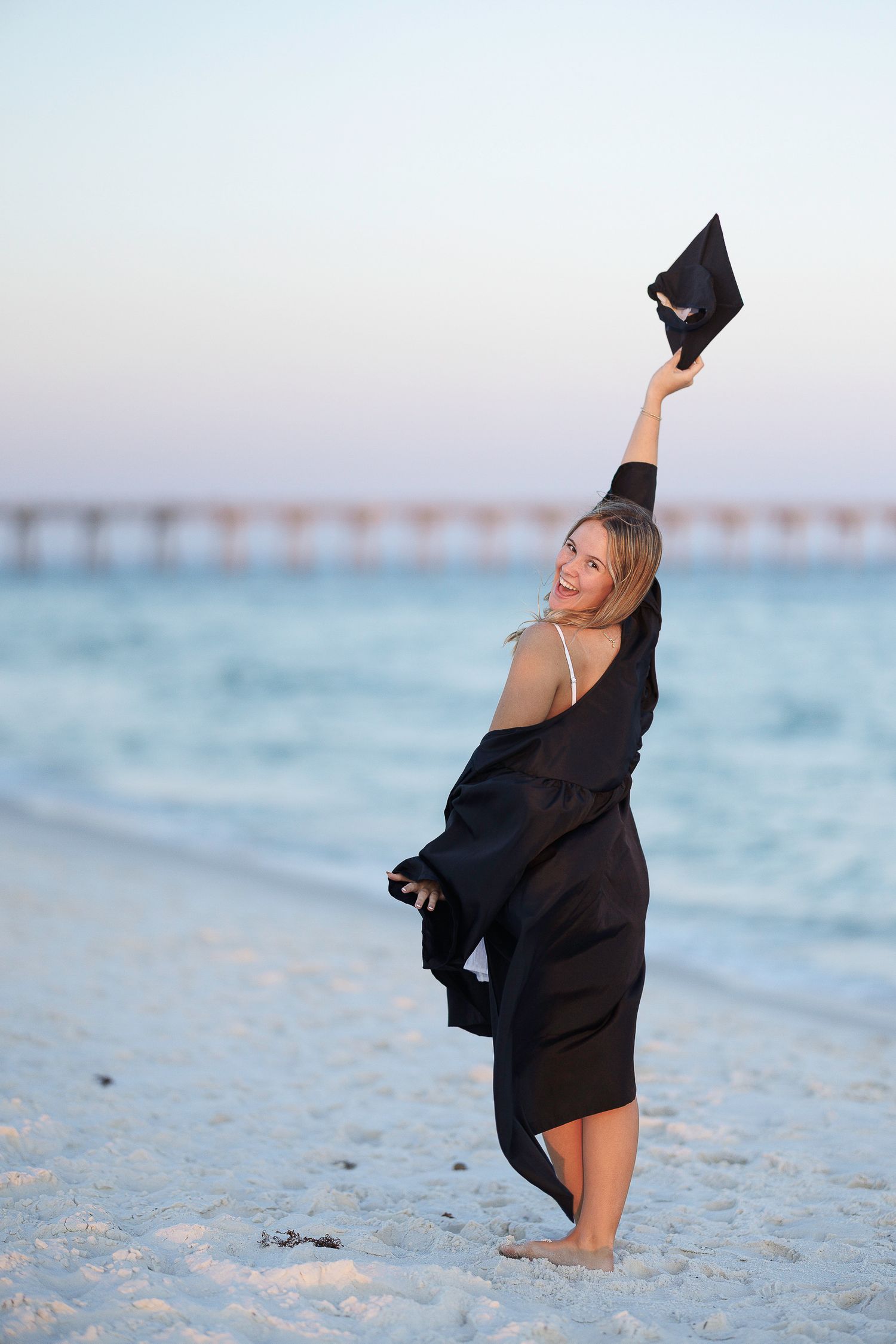 Joyful graduate in black gown raises cap on sandy beach at sunset with pier in background.