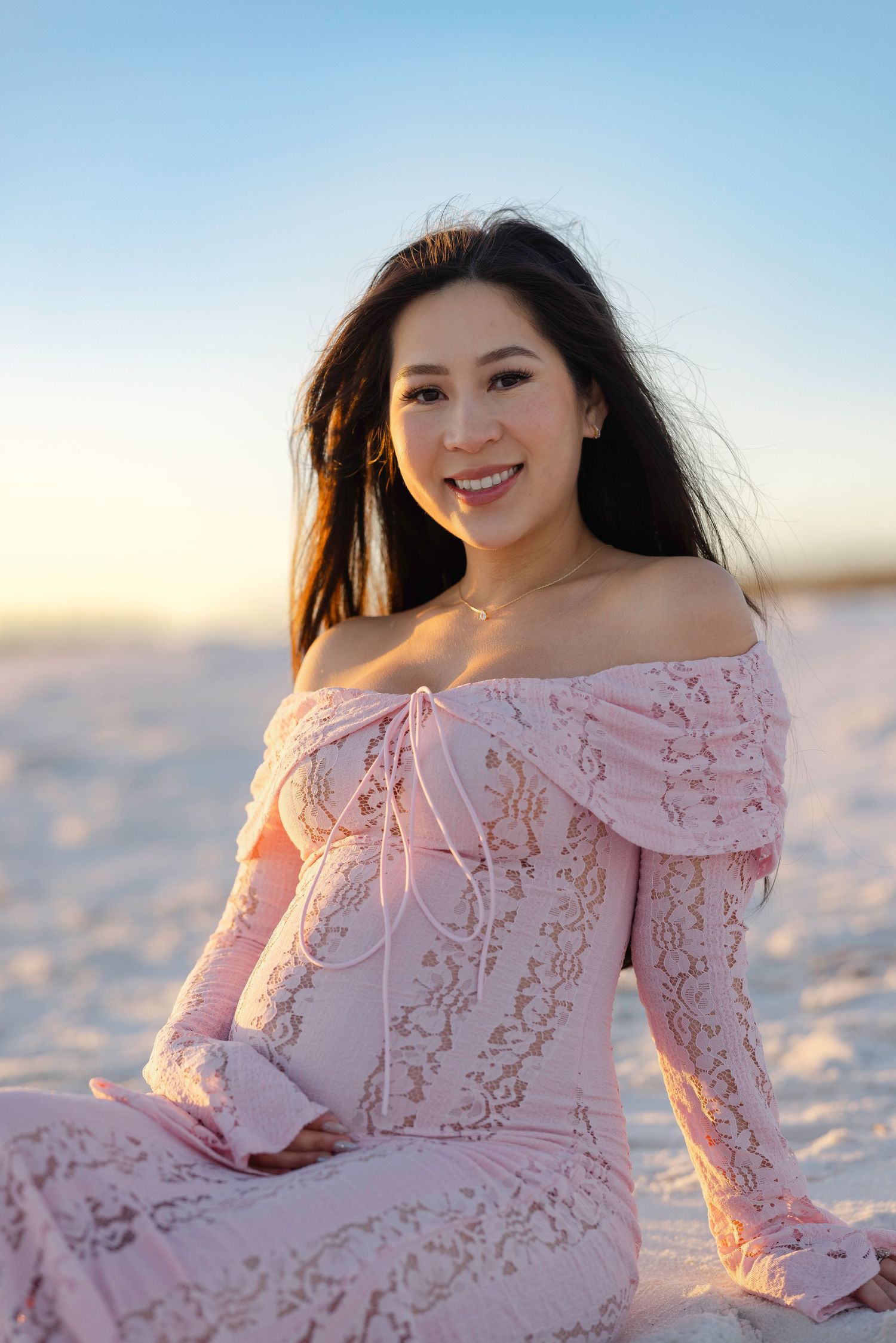 Smiling pregnant Asian woman in pink lace off-shoulder dress sitting on white sand beach at sunset.