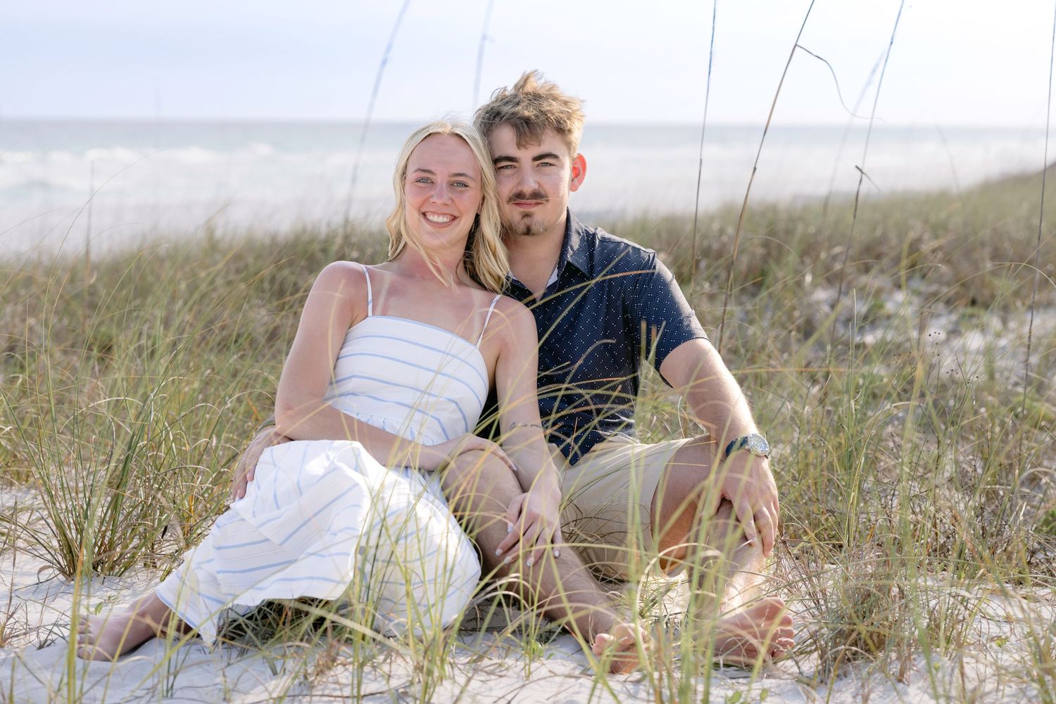 A smiling couple sits together on sandy beach dunes with sea oats, woman in white outfit, man in navy shirt.