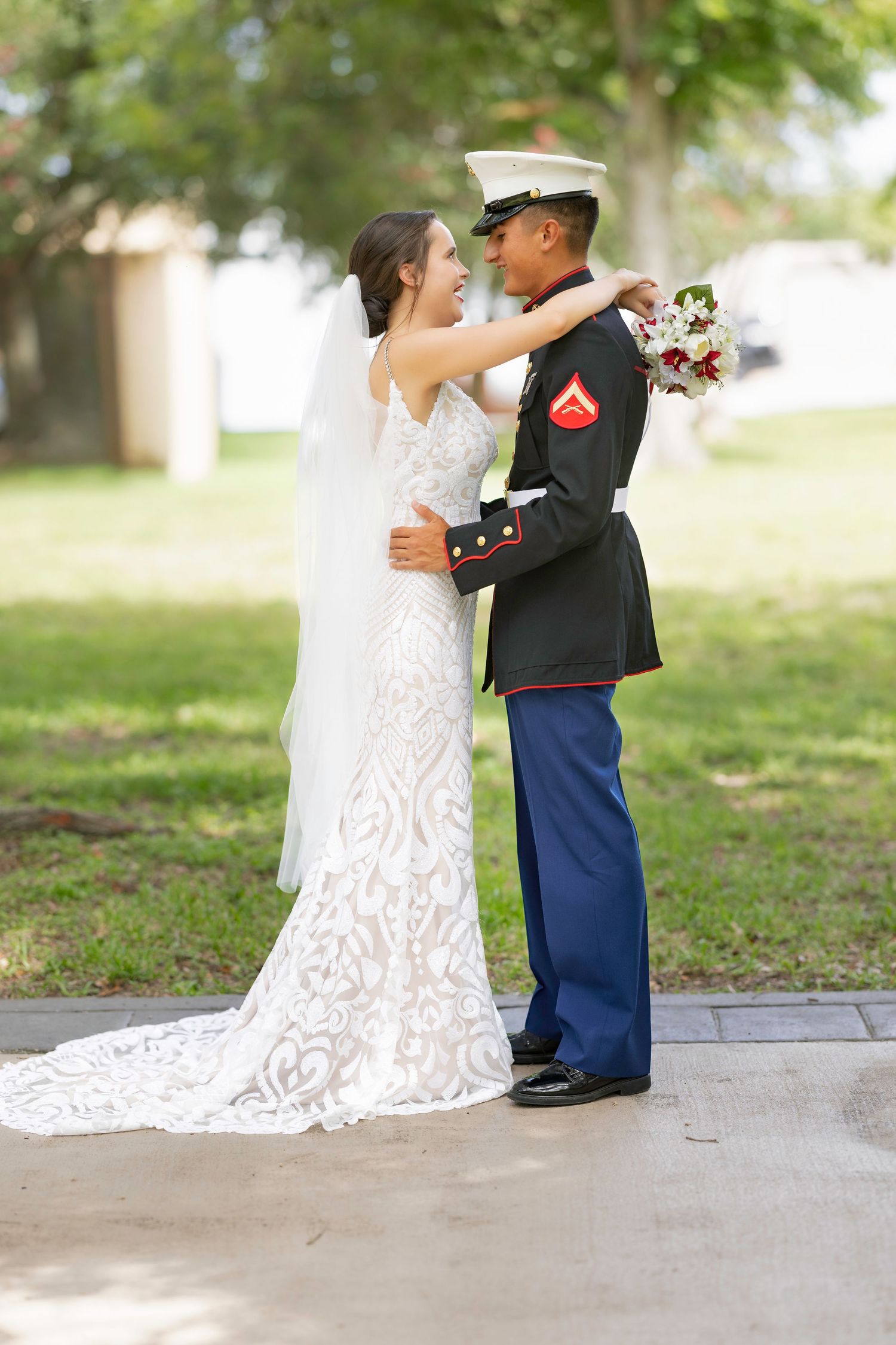 A bride in a lace gown and a Marine in dress uniform embrace outdoors on their wedding day.