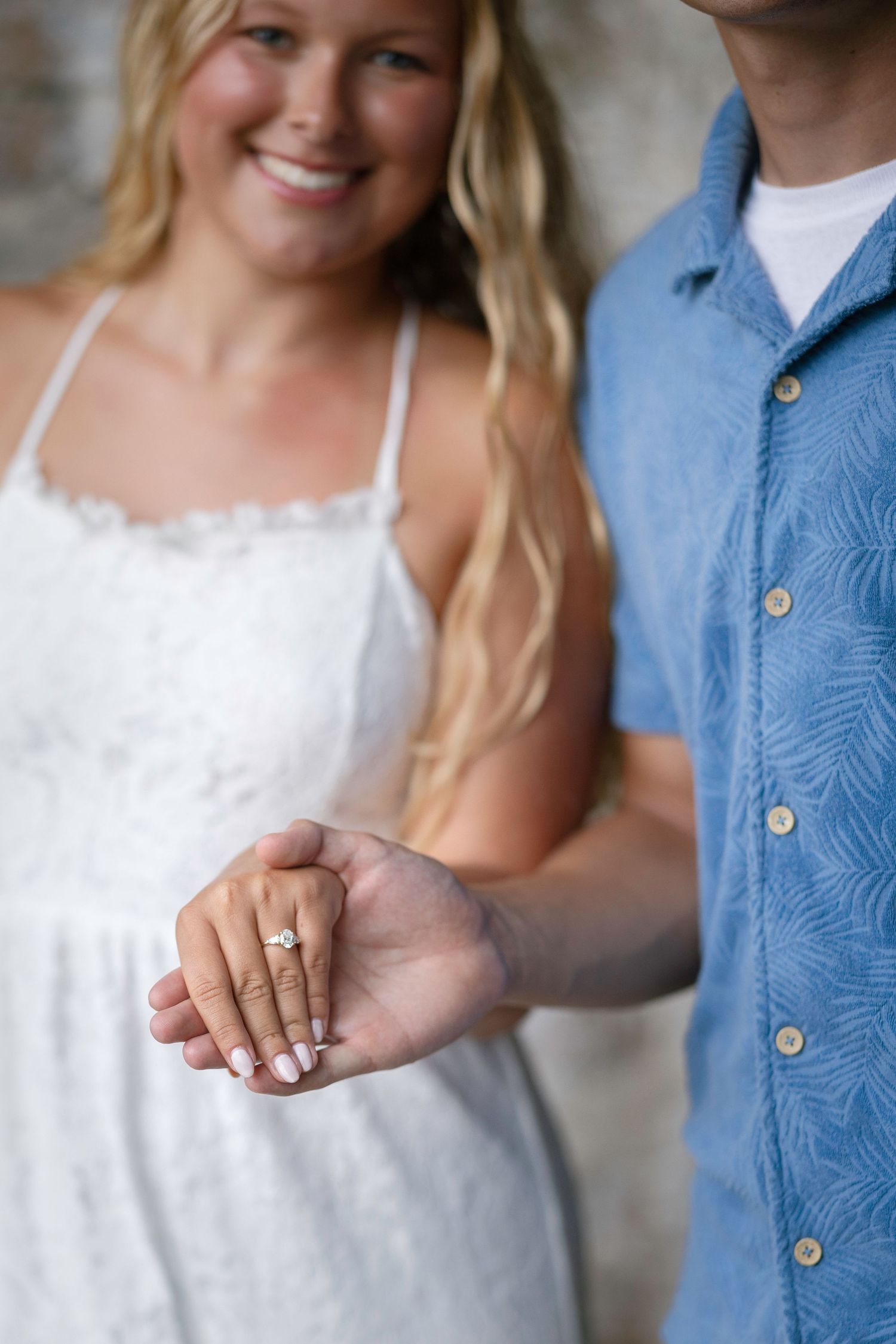 Smiling blonde woman in white dress showing off engagement ring while holding hands with man in blue shirt.