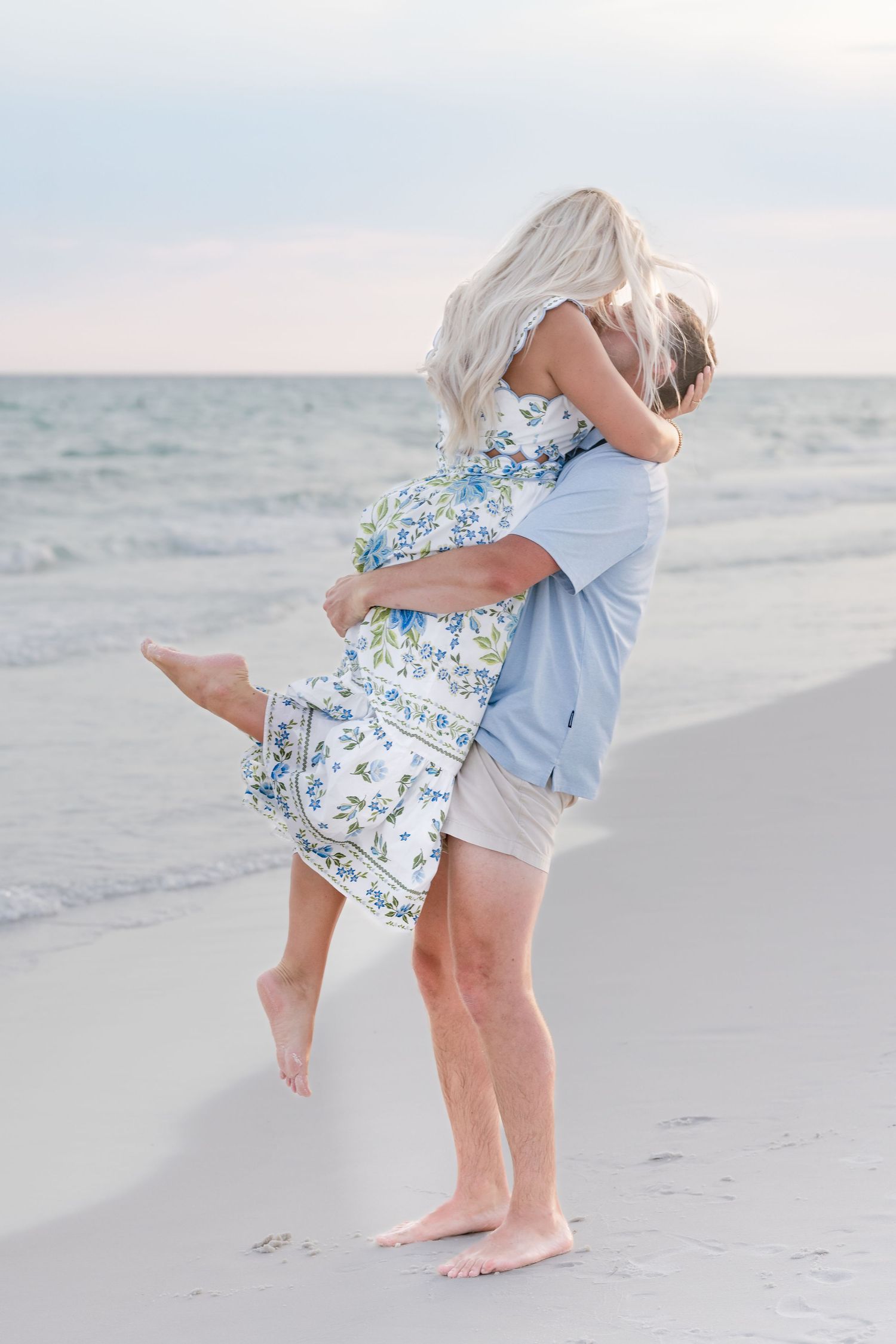 Couple sharing a romantic kiss on a sandy beach at sunset, woman in floral dress being lifted by man in blue shirt.
