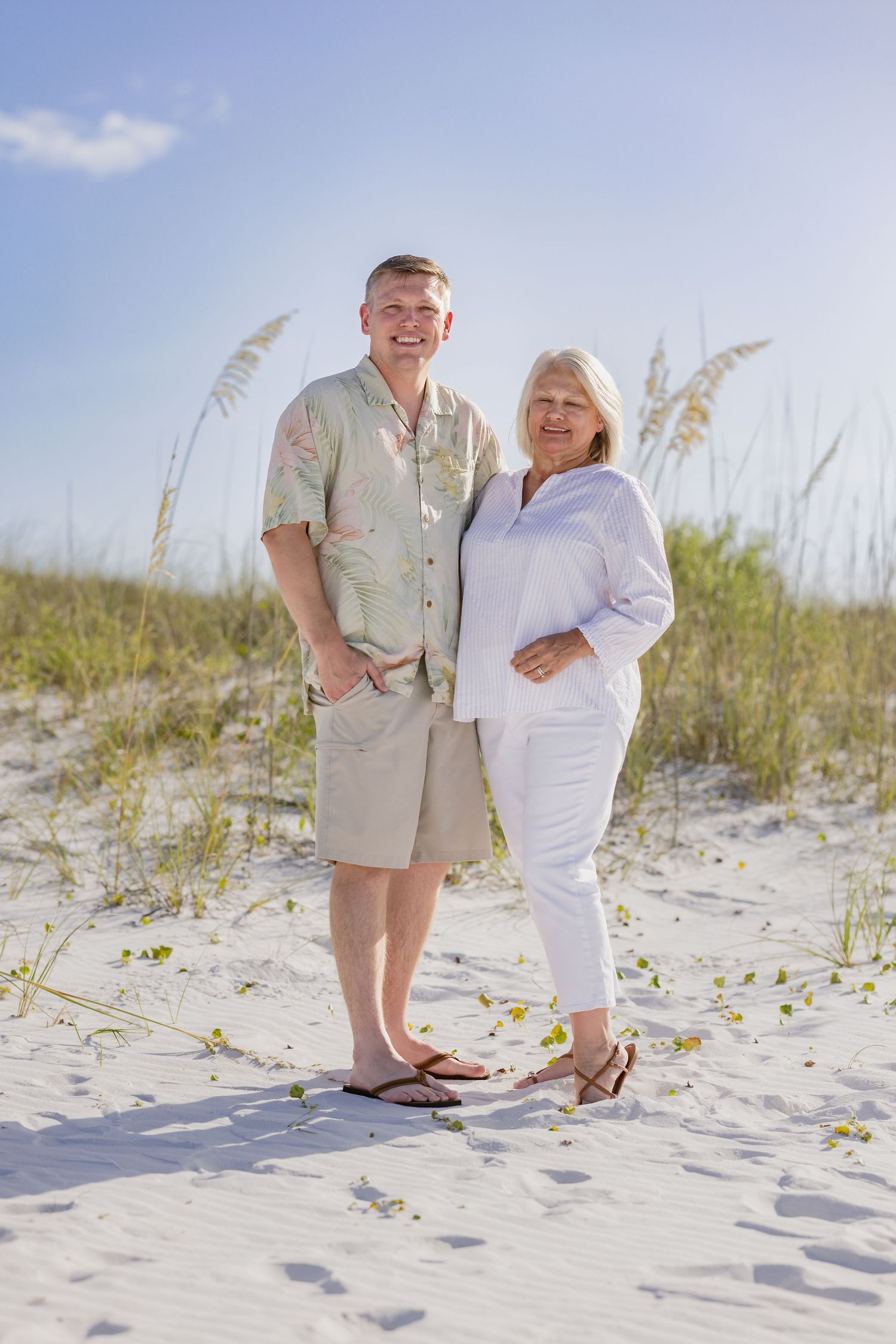 Happy senior couple smiling together on a white sand beach with sea oats and blue sky in background.