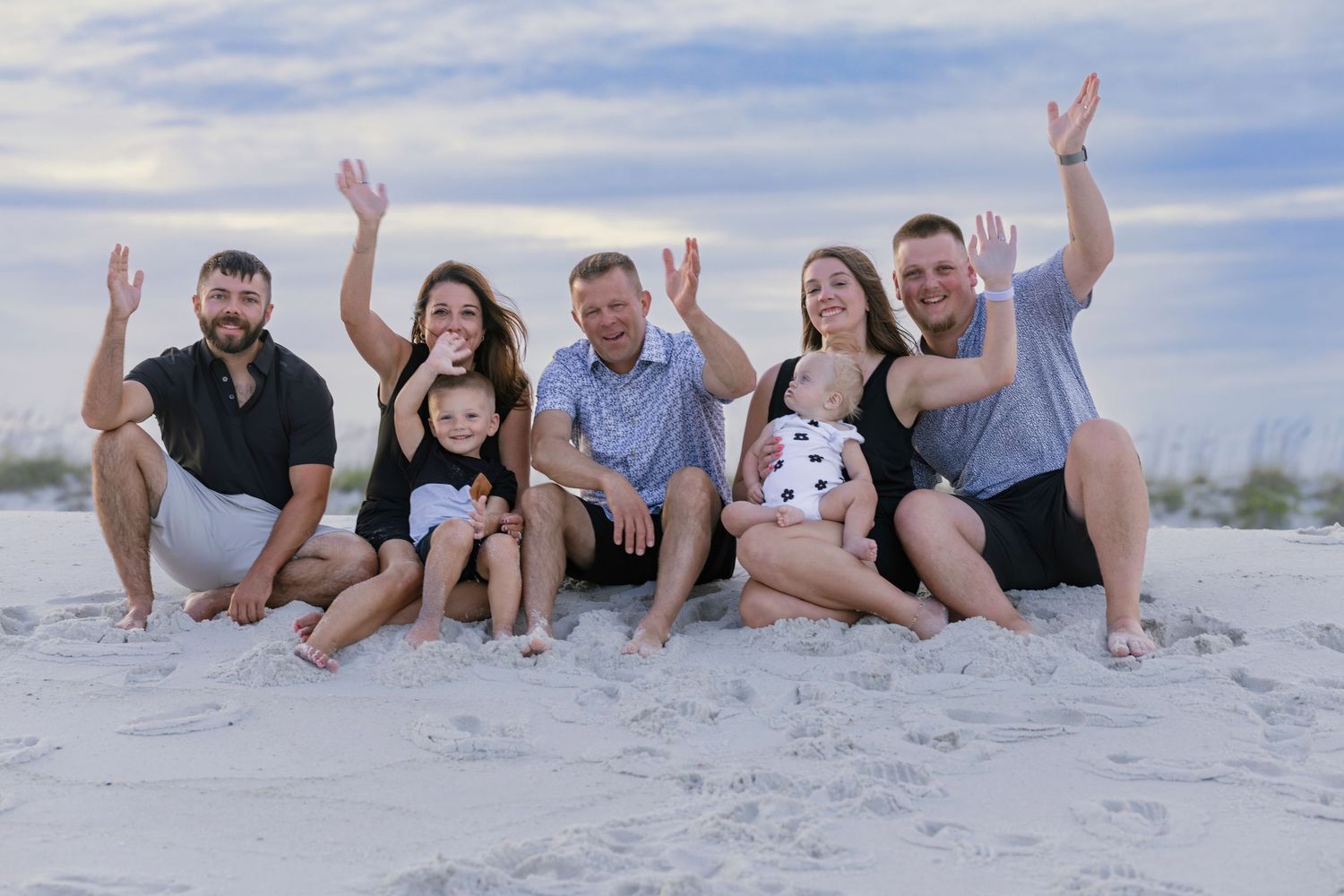 Family of seven posing and waving on a white sand beach with a cloudy sky backdrop.
