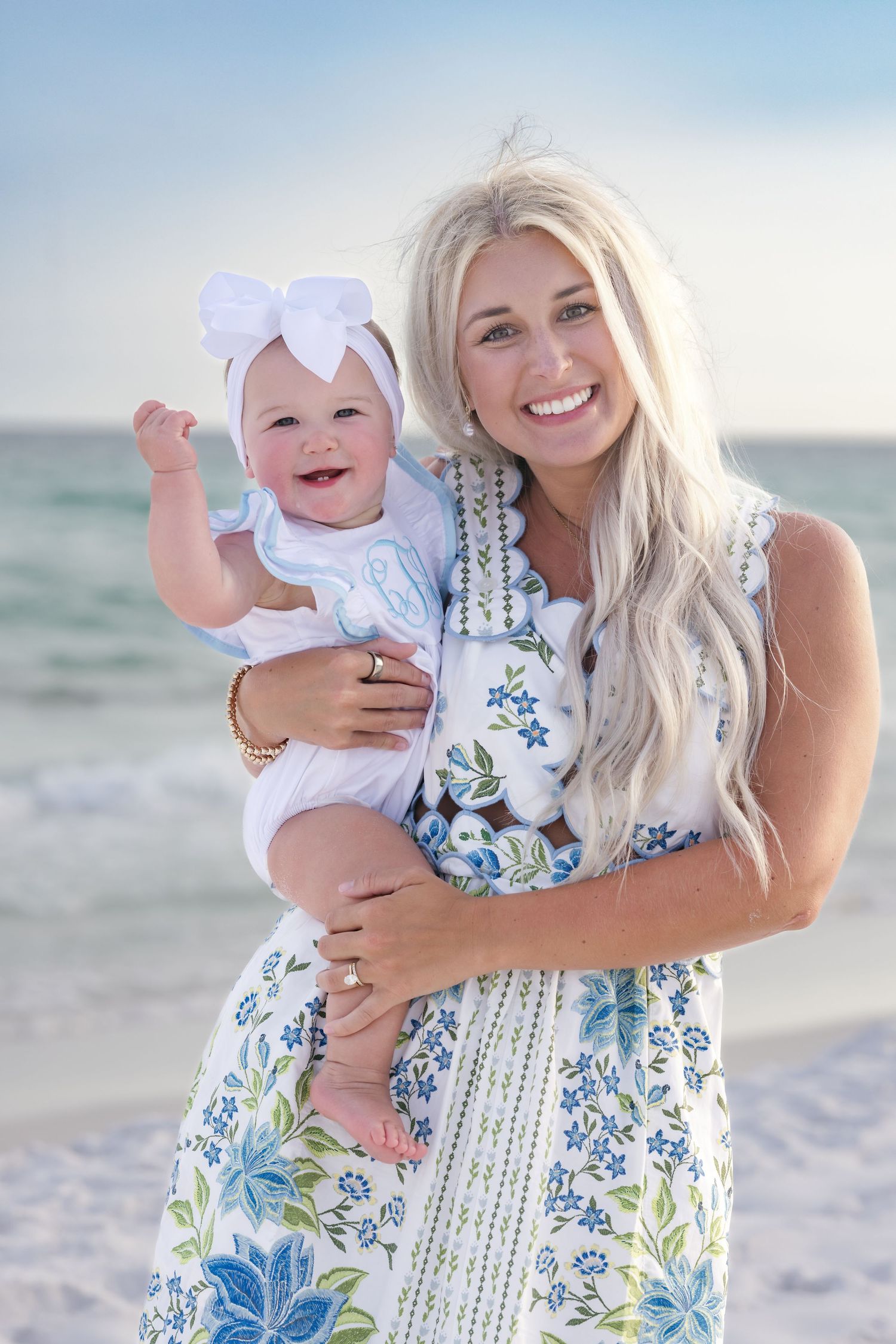 Smiling blonde woman holding a happy baby in a white bow on a sunny beach, both wearing blue floral outfits.