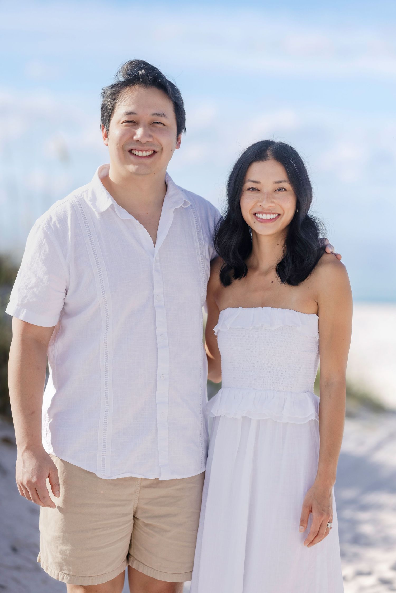 Smiling couple in white outfits posing together on a sunny beach with blue skies.