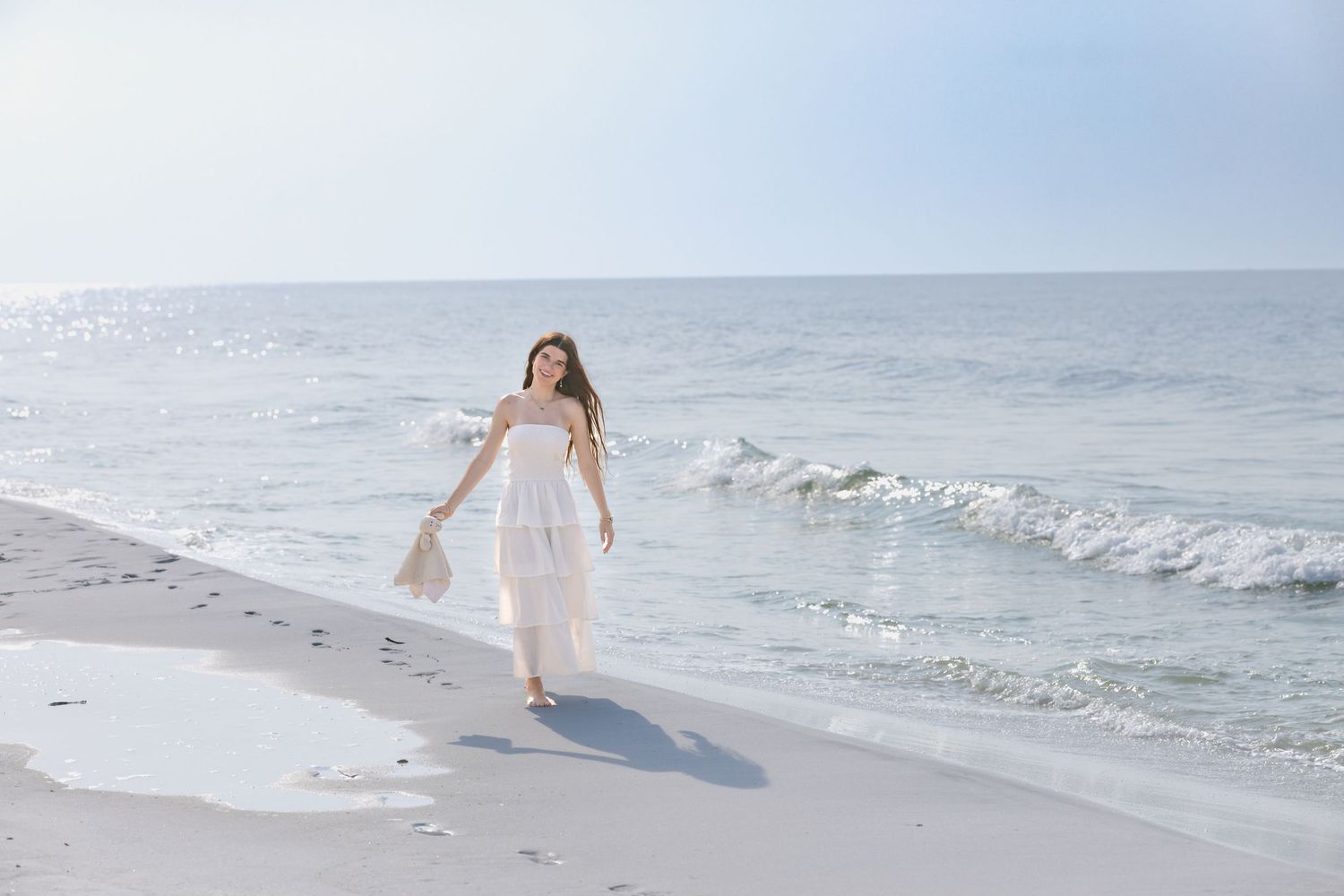 Woman in white strapless dress walking along a sunny beach carrying a bag near ocean waves.
