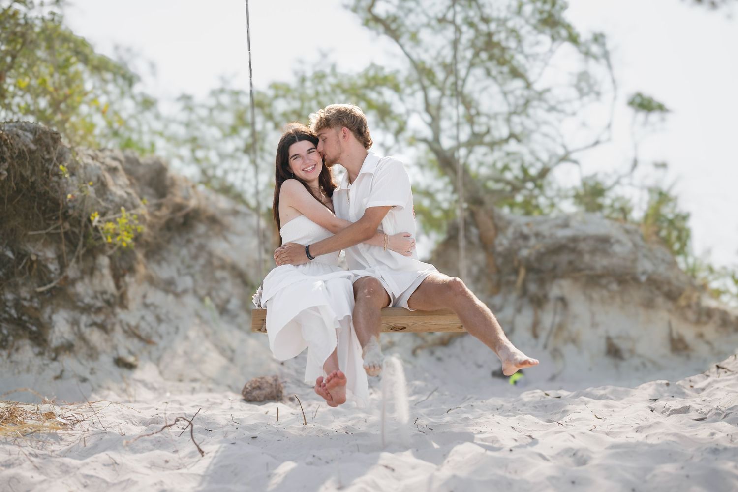 Romantic couple sharing a tender kiss on a rope swing on a sandy beach, dressed in white outfits.