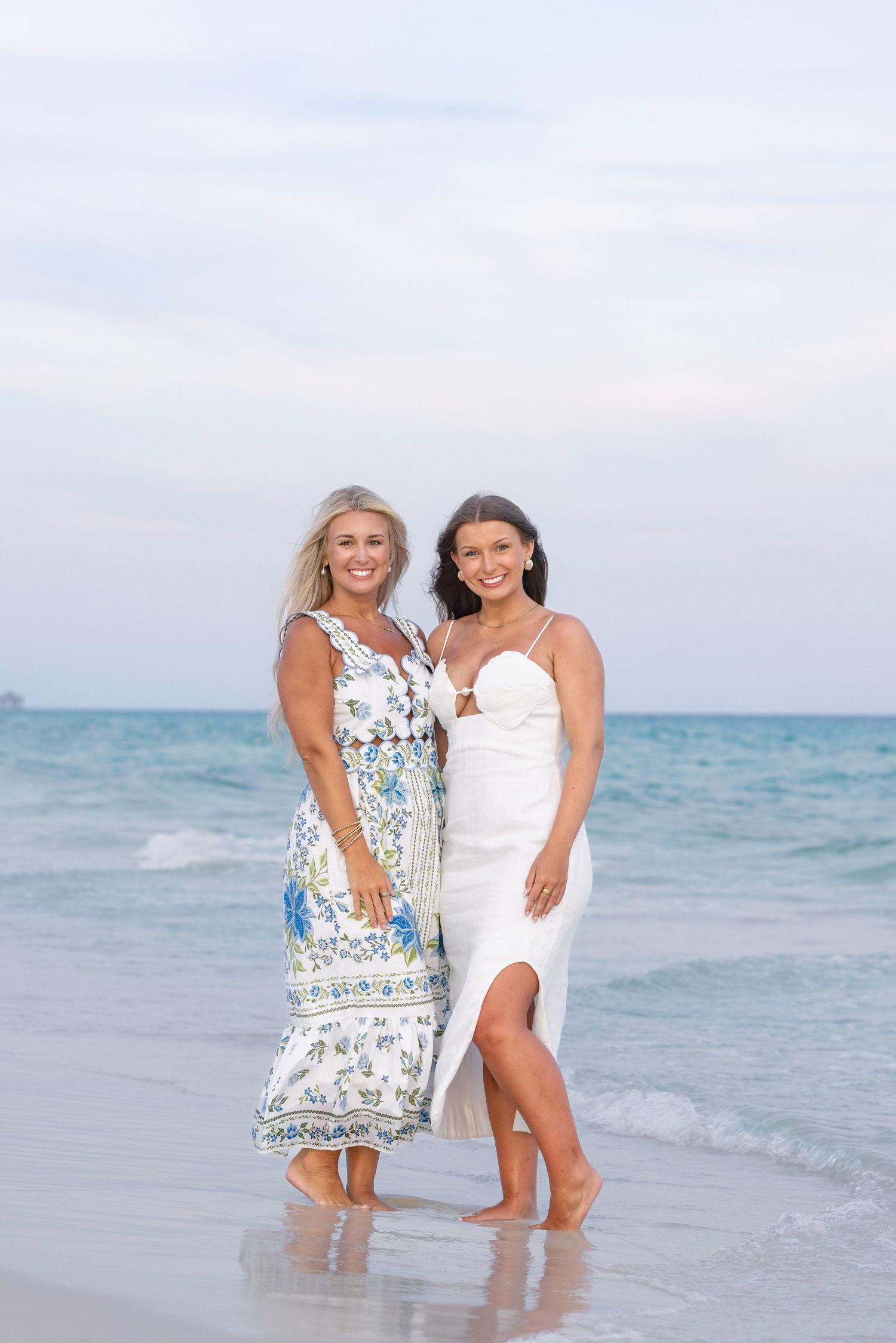 Two smiling women in summer dresses standing together on sandy beach with ocean behind them.