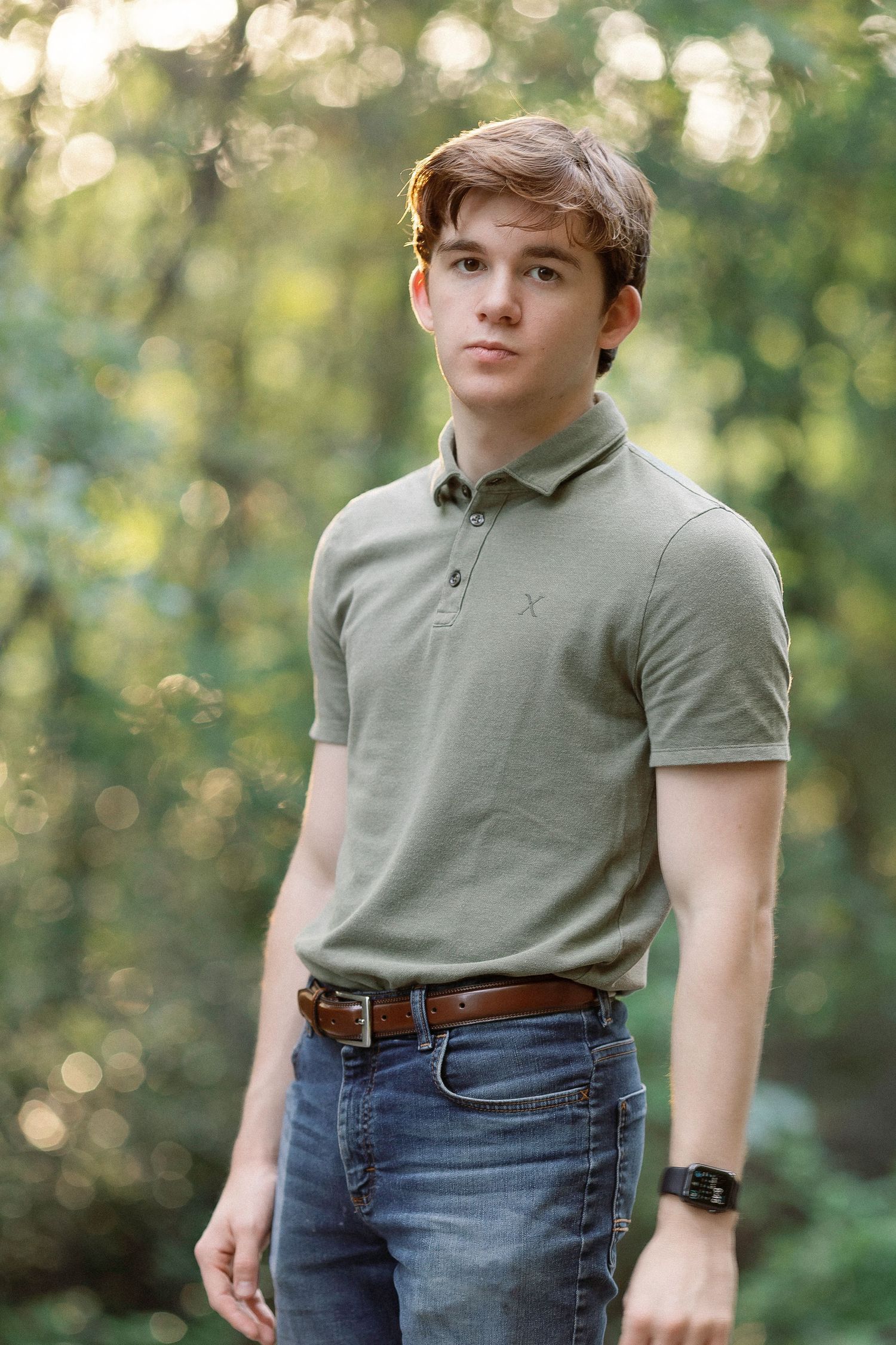 Young man in olive polo shirt and jeans poses outdoors in lush green forest setting.