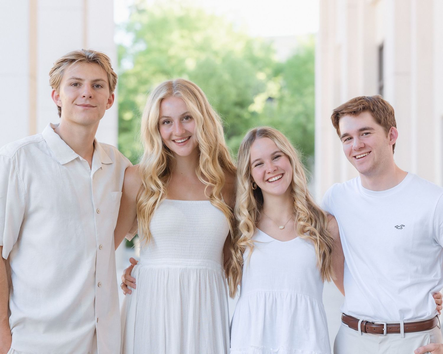 Four smiling young adults posing together in white and cream coordinating outfits outdoors.