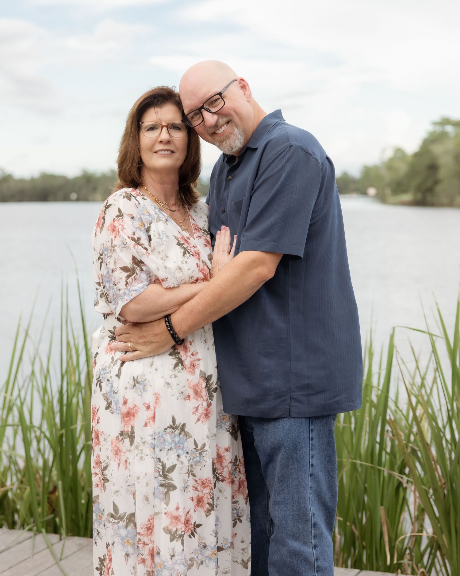 Couple embracing by a serene lake, woman in floral dress and man in navy shirt, sharing a tender moment outdoors.