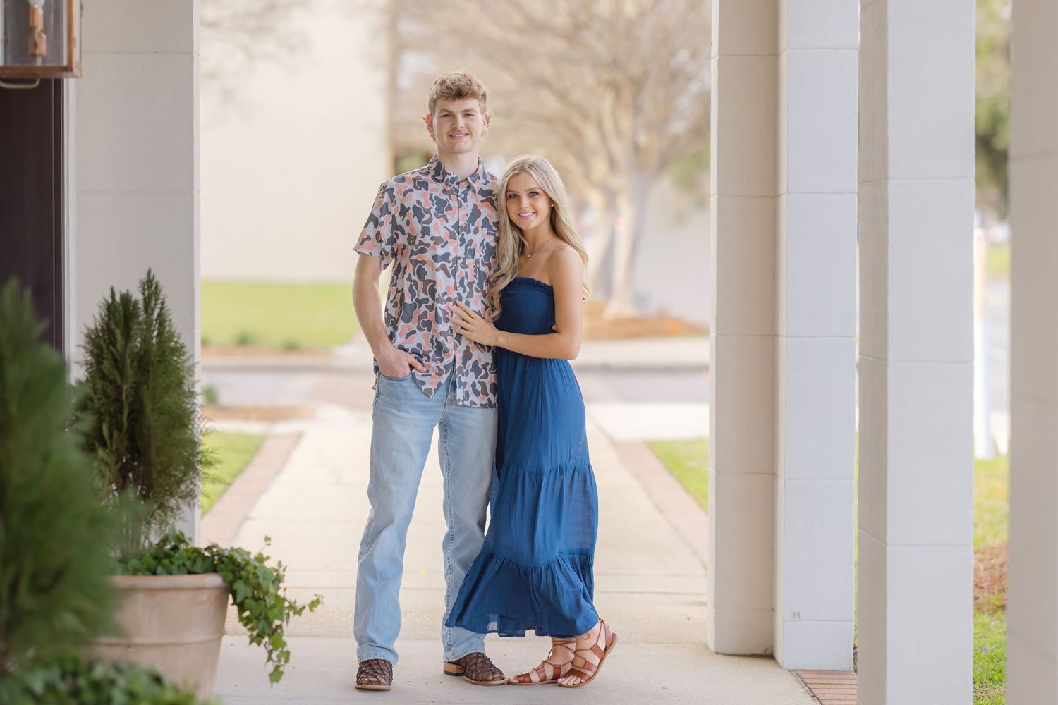 A couple in blue dress and jeans poses together on a white columned porch during a sunny day.