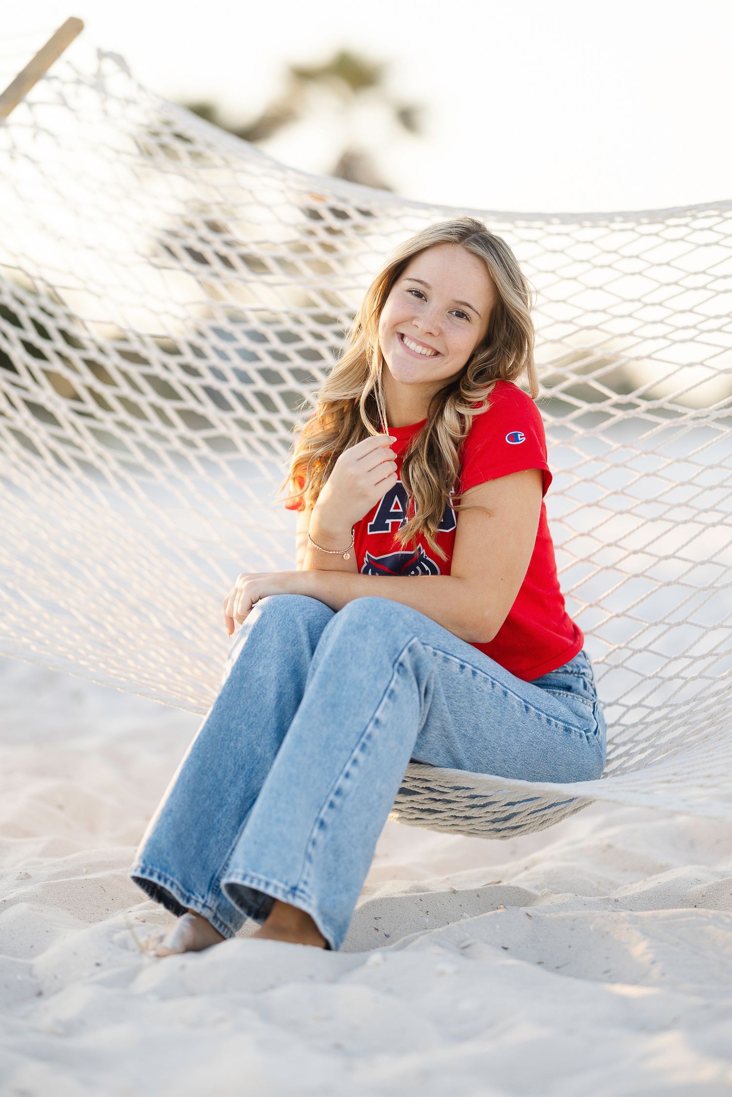 Person in red top and jeans relaxing in a hammock on a sandy beach during sunset.