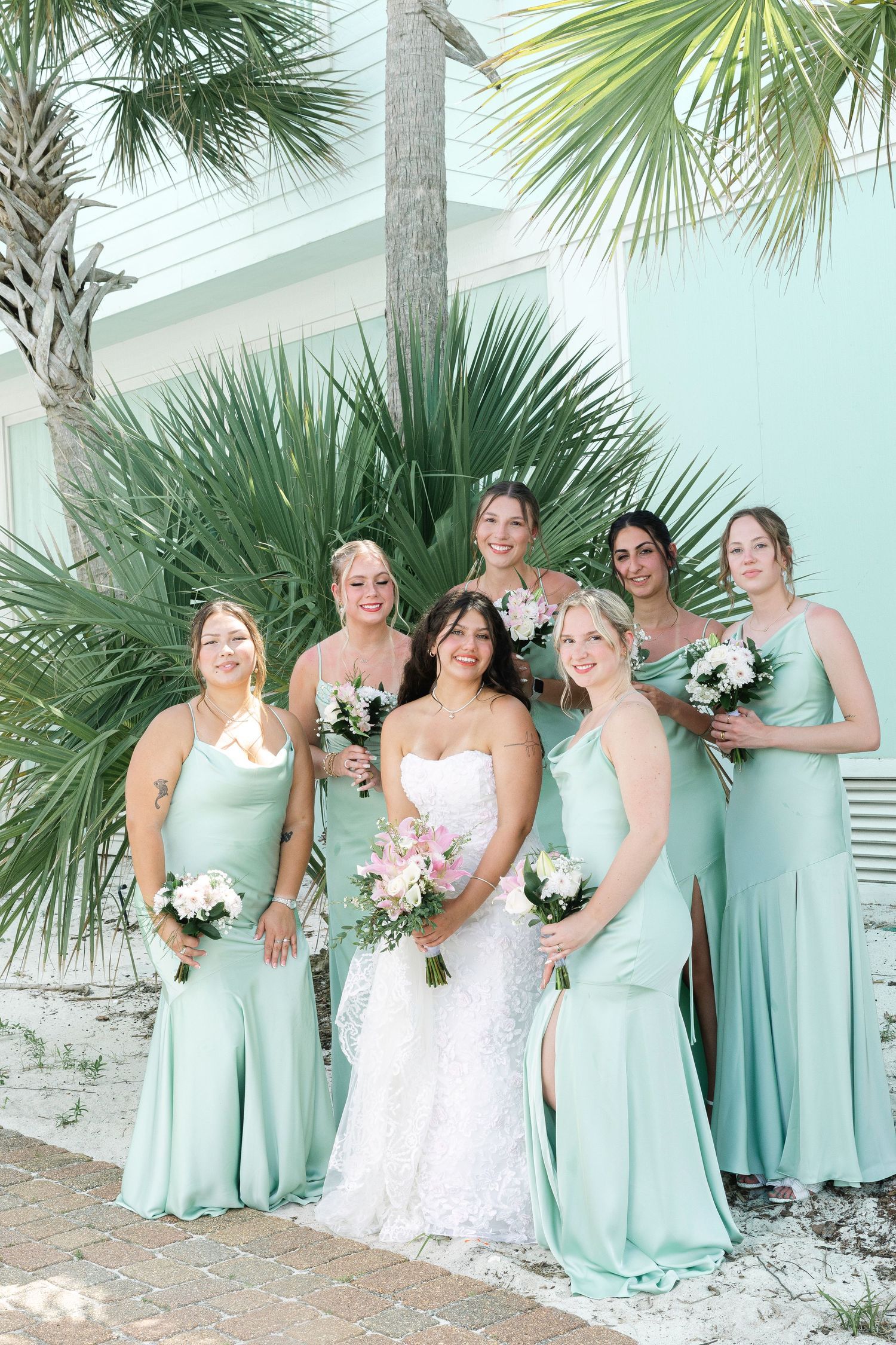Wedding party in mint green dresses and white gown poses against palm trees at a beachfront venue.