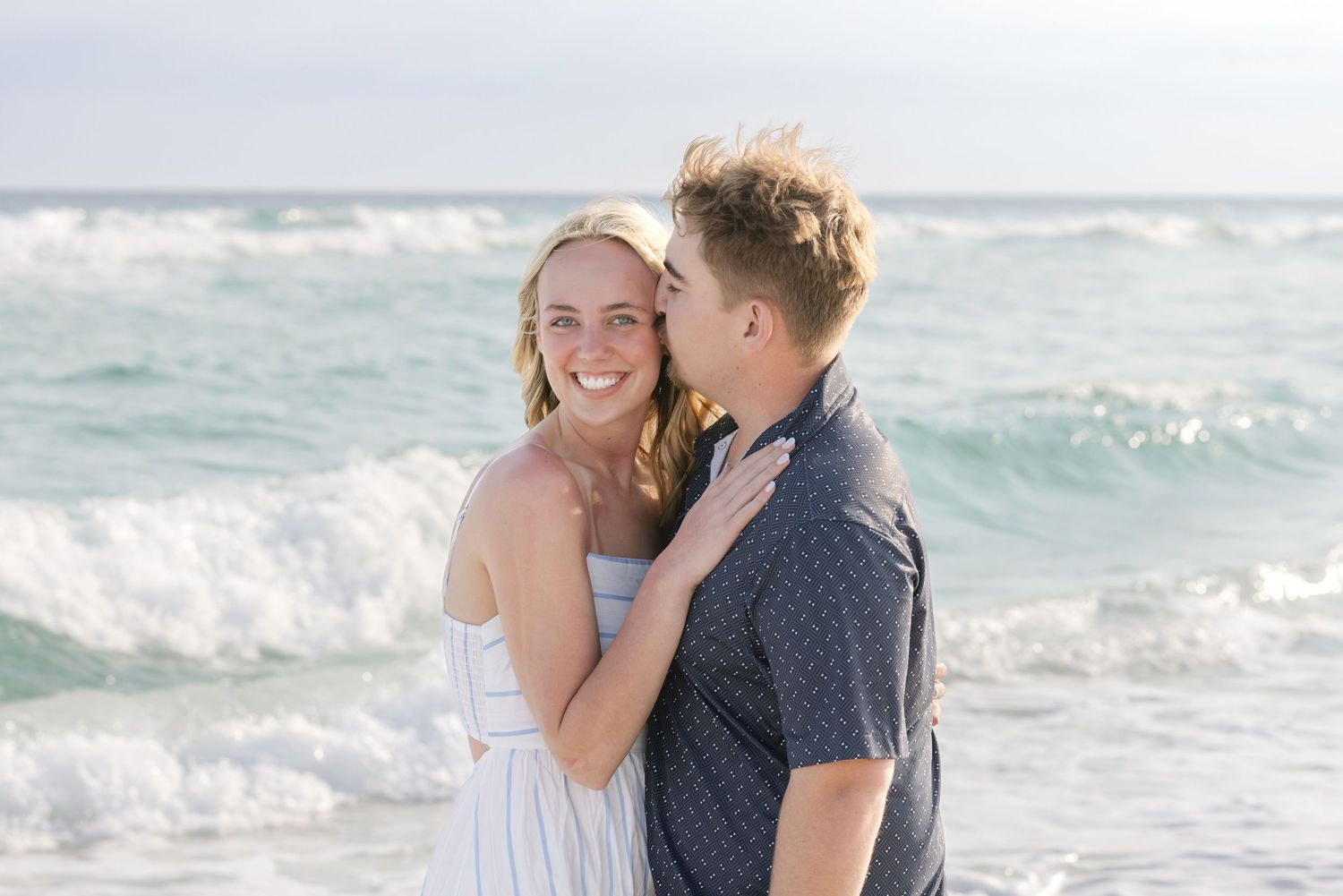 A smiling couple embraces on a sunny beach with turquoise ocean waves behind them.