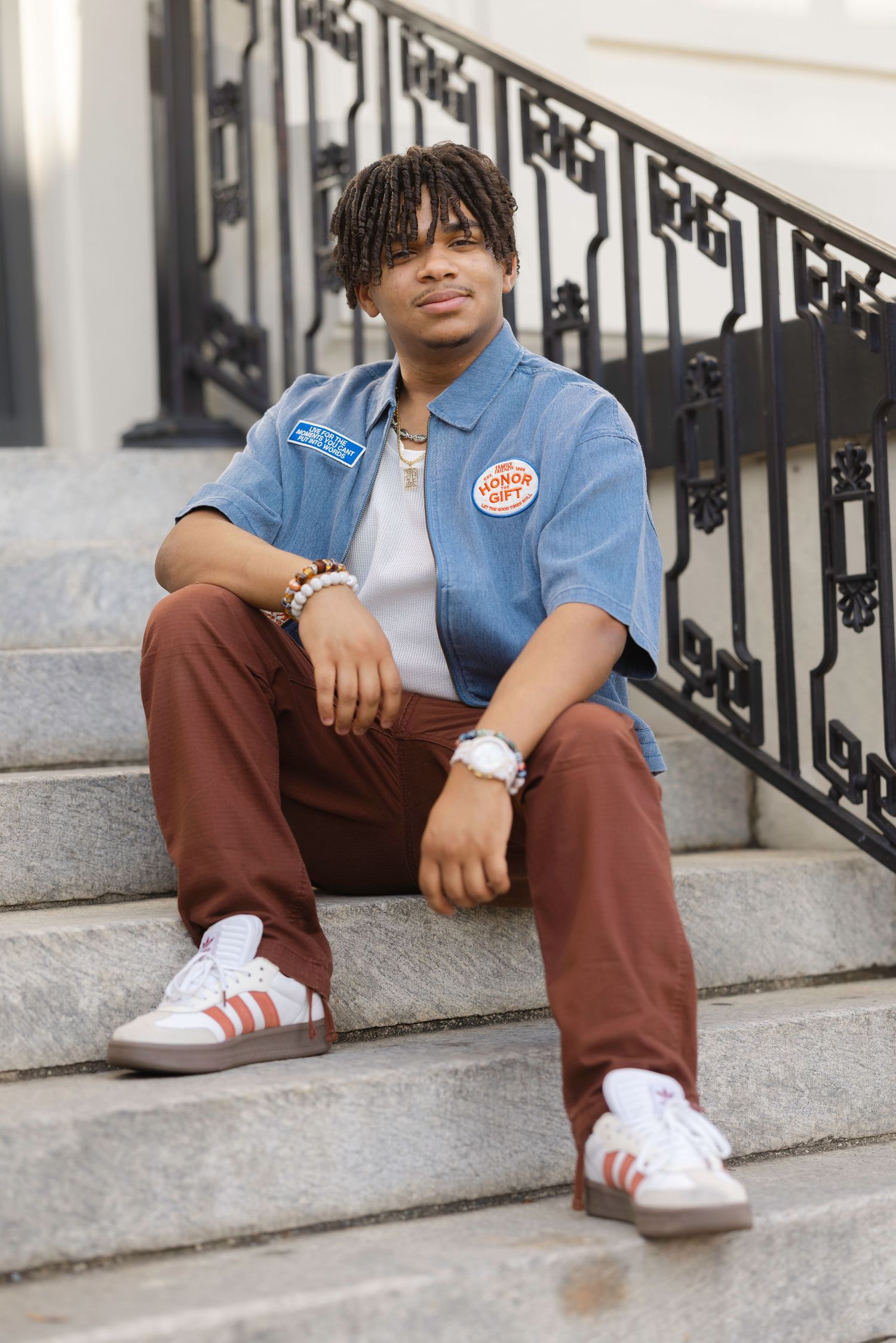 Young man in blue shirt, brown pants, and Adidas sneakers sitting on stone steps with iron railing.