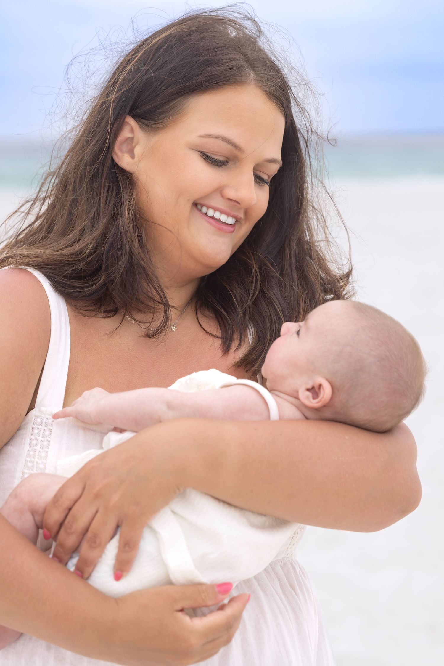 Smiling mother in white dress holding newborn baby on a sunny beach.
