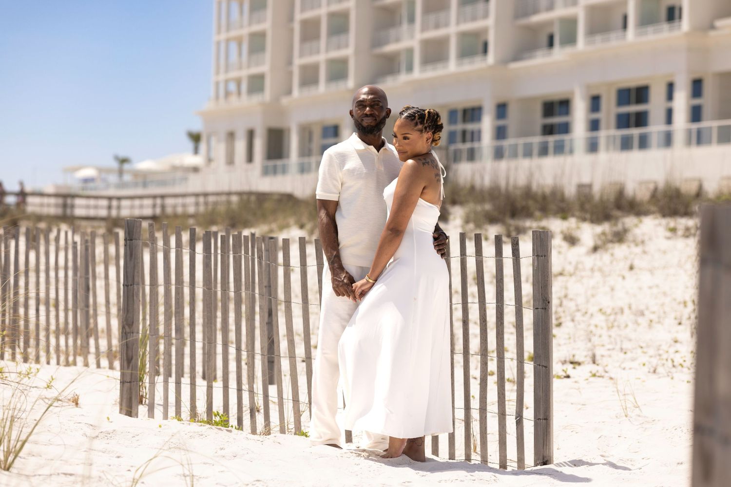 A couple in white attire poses romantically on a sandy beach with a wooden fence and resort building backdrop.