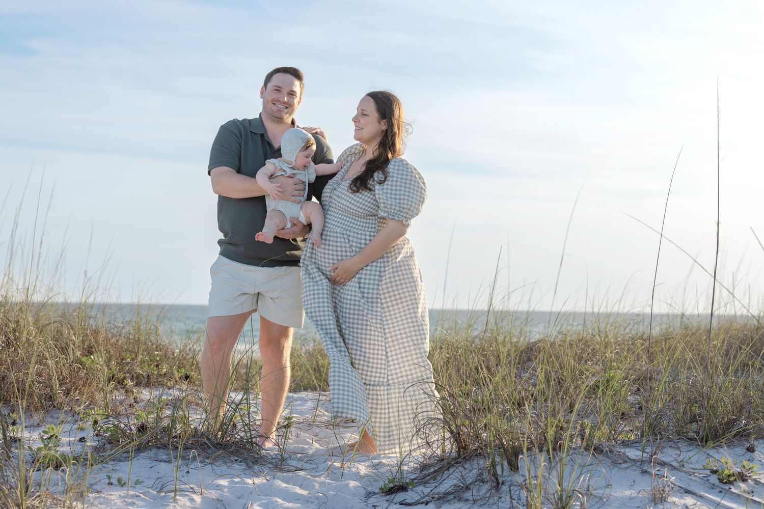 Pregnant woman and man holding baby pose on sandy beach with sea oats and ocean in background.