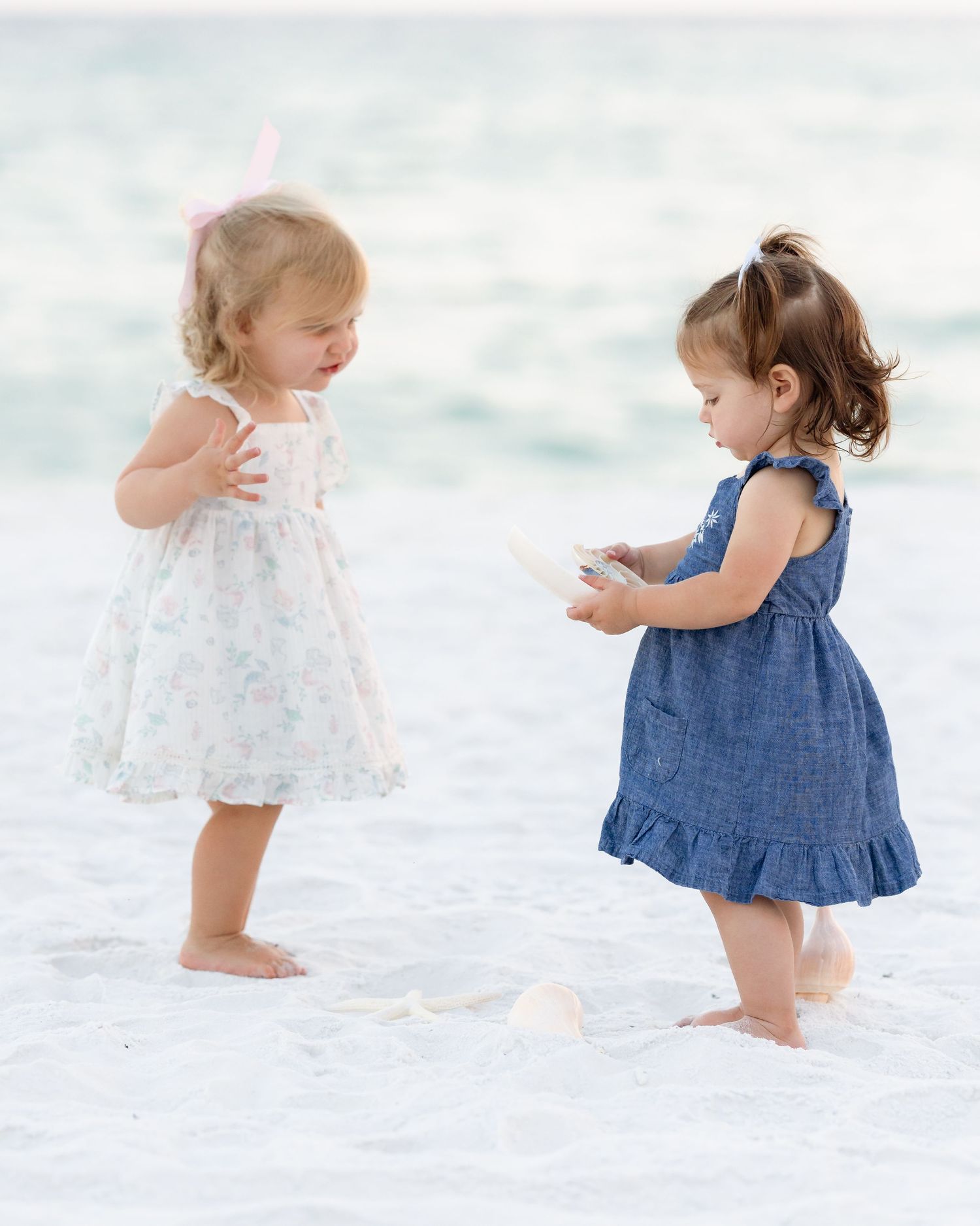 Two toddler girls in summer dresses play together on a sandy beach near the ocean shoreline.