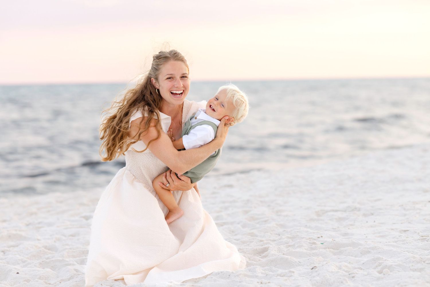 Five candid photos show a couple in white outfits embracing on a sunlit beach with gentle waves breaking behind them.
