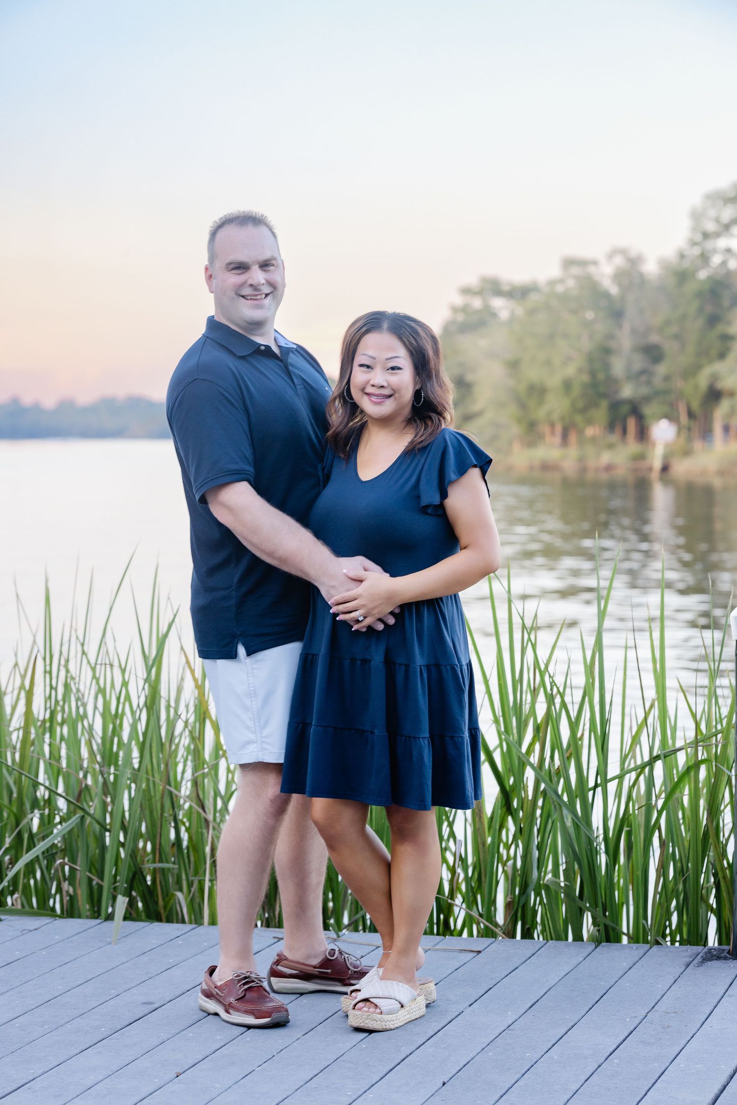 Couple in matching navy outfits smiling on a lakeside dock at sunset, surrounded by tall green reeds.