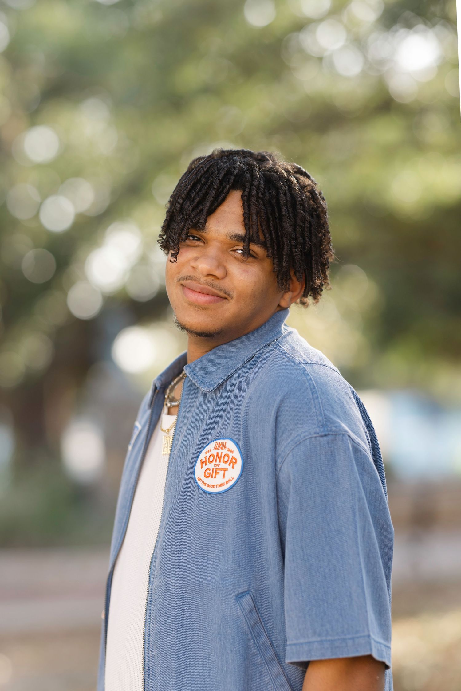 Young man with braids wearing a blue shirt with a badge, smiling outdoors with blurred trees in background.