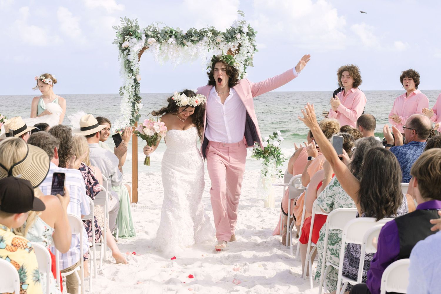 Joyful couple walks down beach wedding aisle, groom in pink suit, bride in white dress, guests cheering.