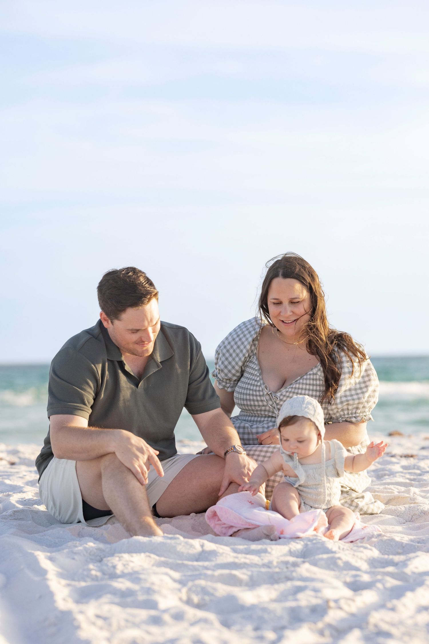 A young family sits on white sand beach, parents smiling at their baby in a pink outfit and bonnet.