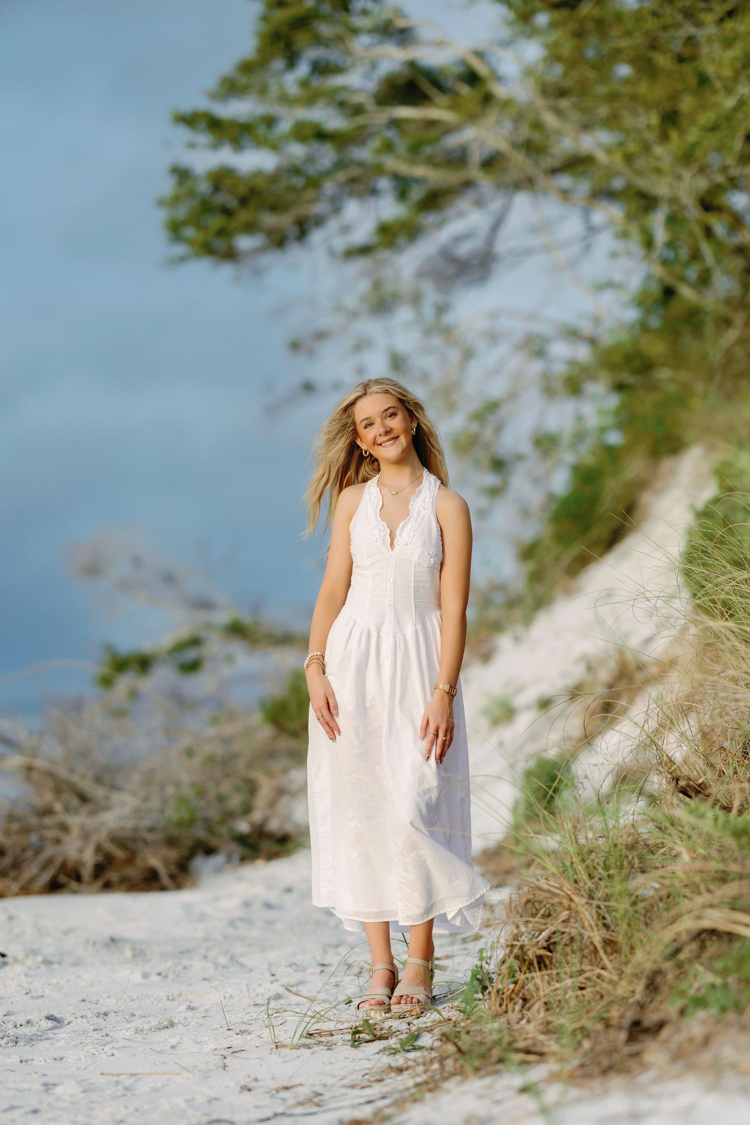 Young blonde woman in white midi dress smiling on sandy beach path with trees and blue sky background.