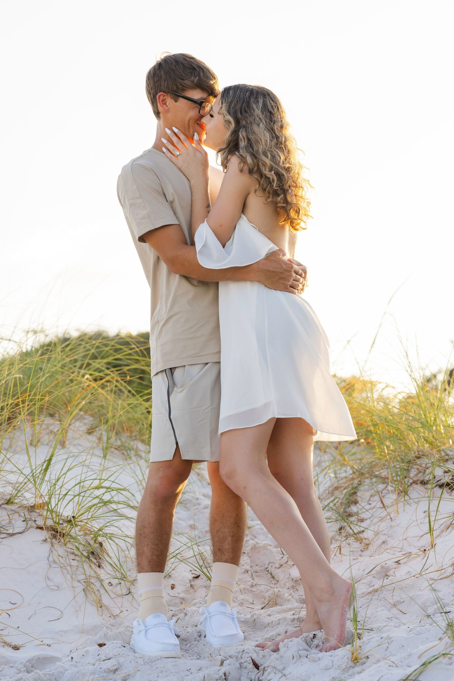 Romantic couple embracing and kissing on sandy beach dunes at sunset, woman in white dress, man in beige outfit.