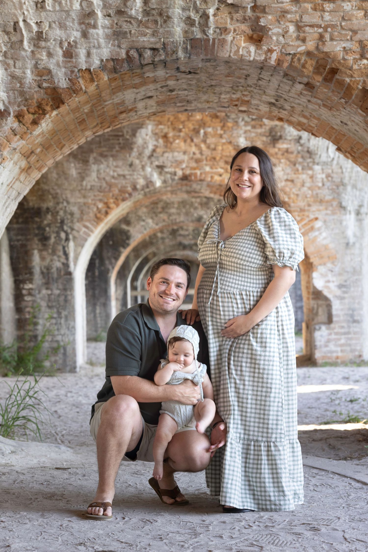 Family portrait under brick arches, pregnant woman in plaid dress, man kneeling holding baby, historic fort setting.