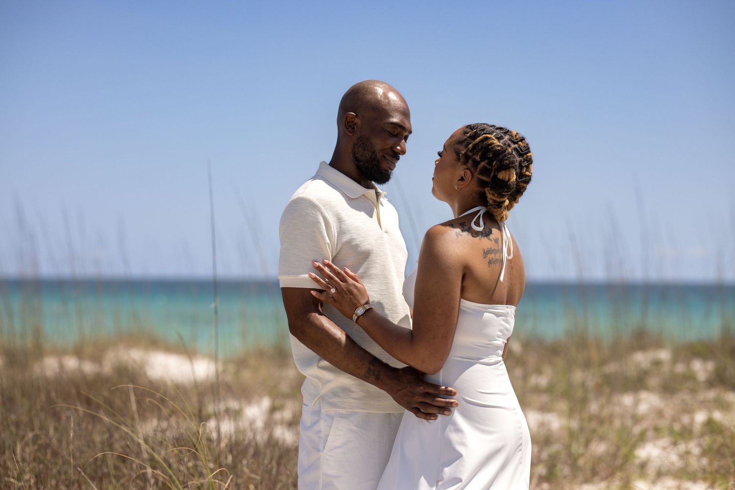Couple in white attire embracing on a sandy beach with turquoise ocean waters in the background.