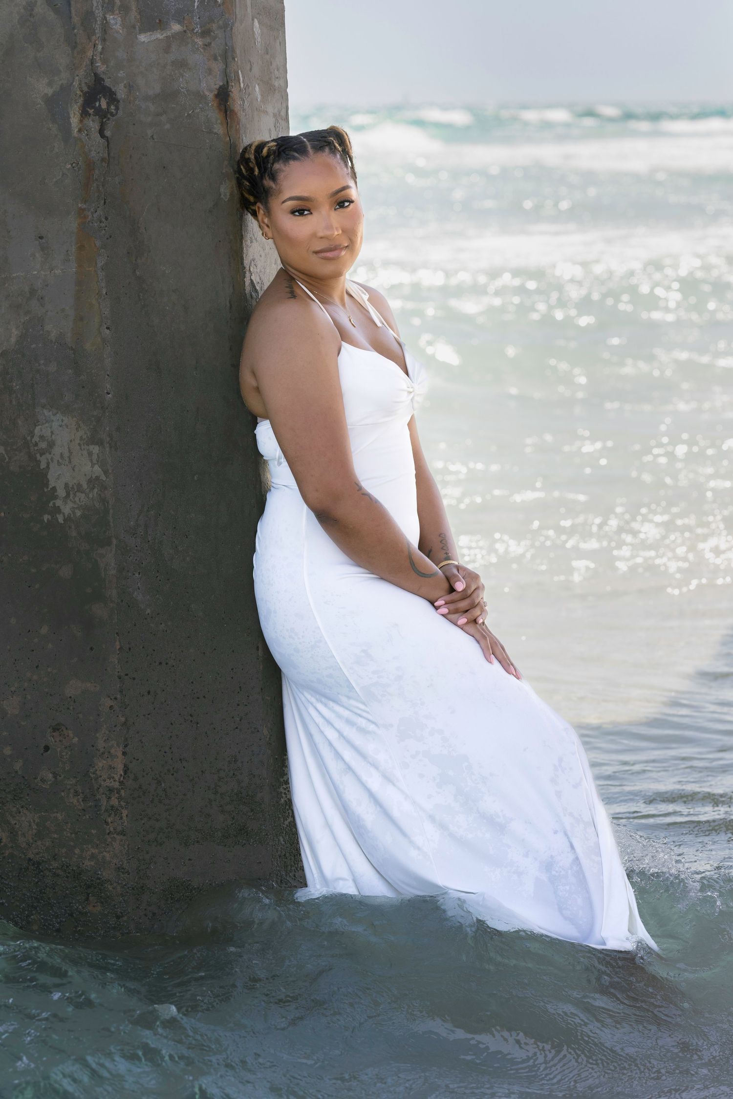 Woman in white gown leaning against pier pillar in ocean waves at beach.