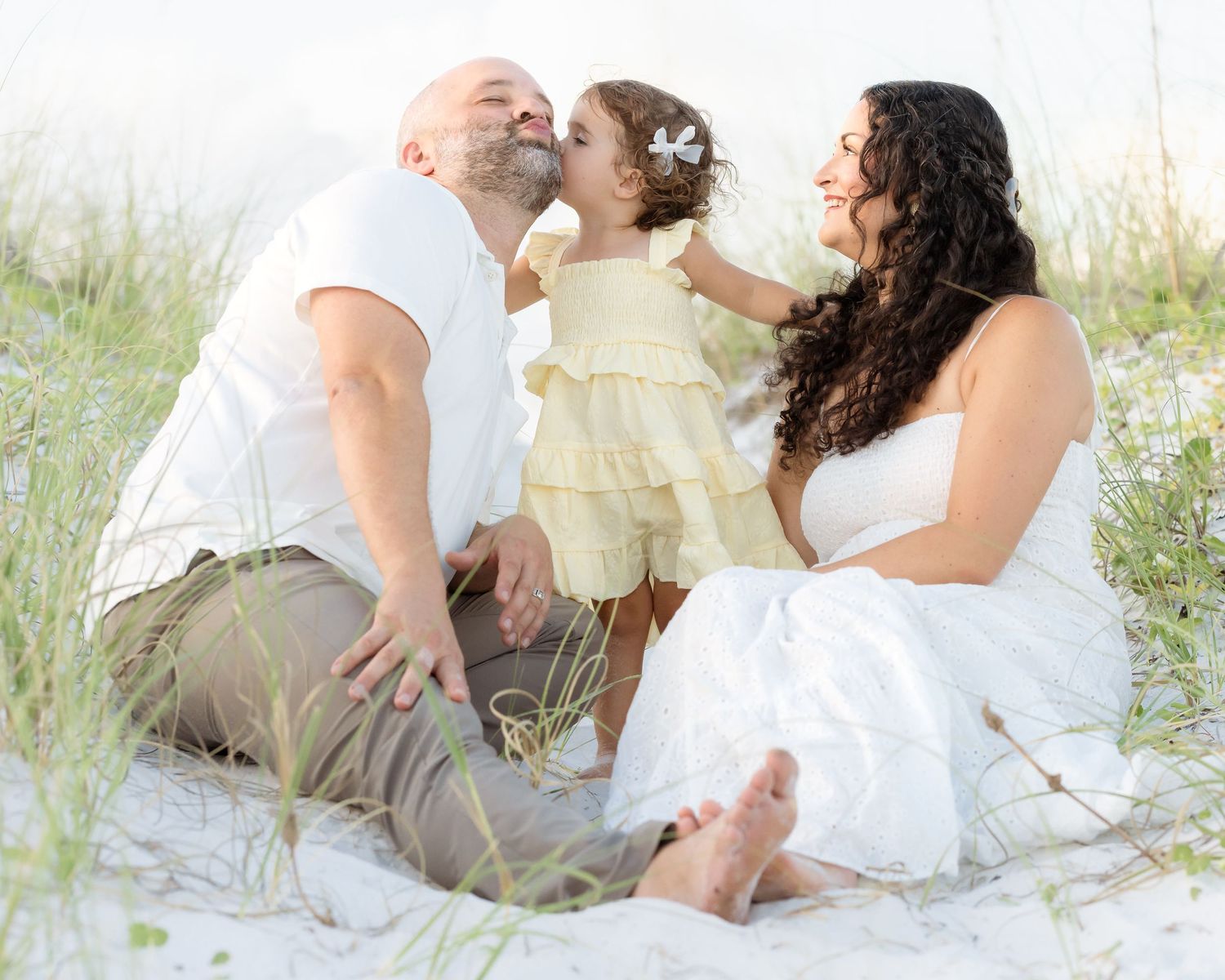 Family beach photo with parents kissing toddler girl in yellow dress while sitting on white sand dunes.