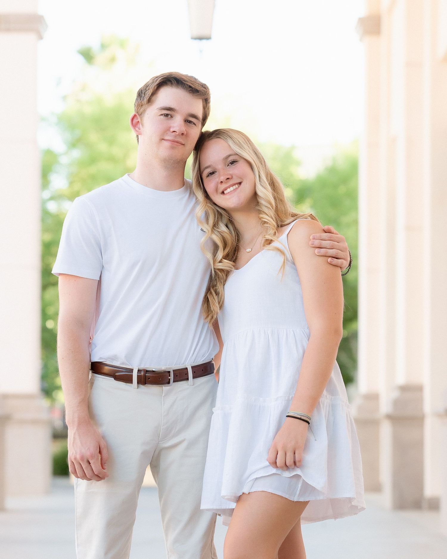 A young couple in matching white outfits poses smiling between elegant columns in an outdoor portrait.