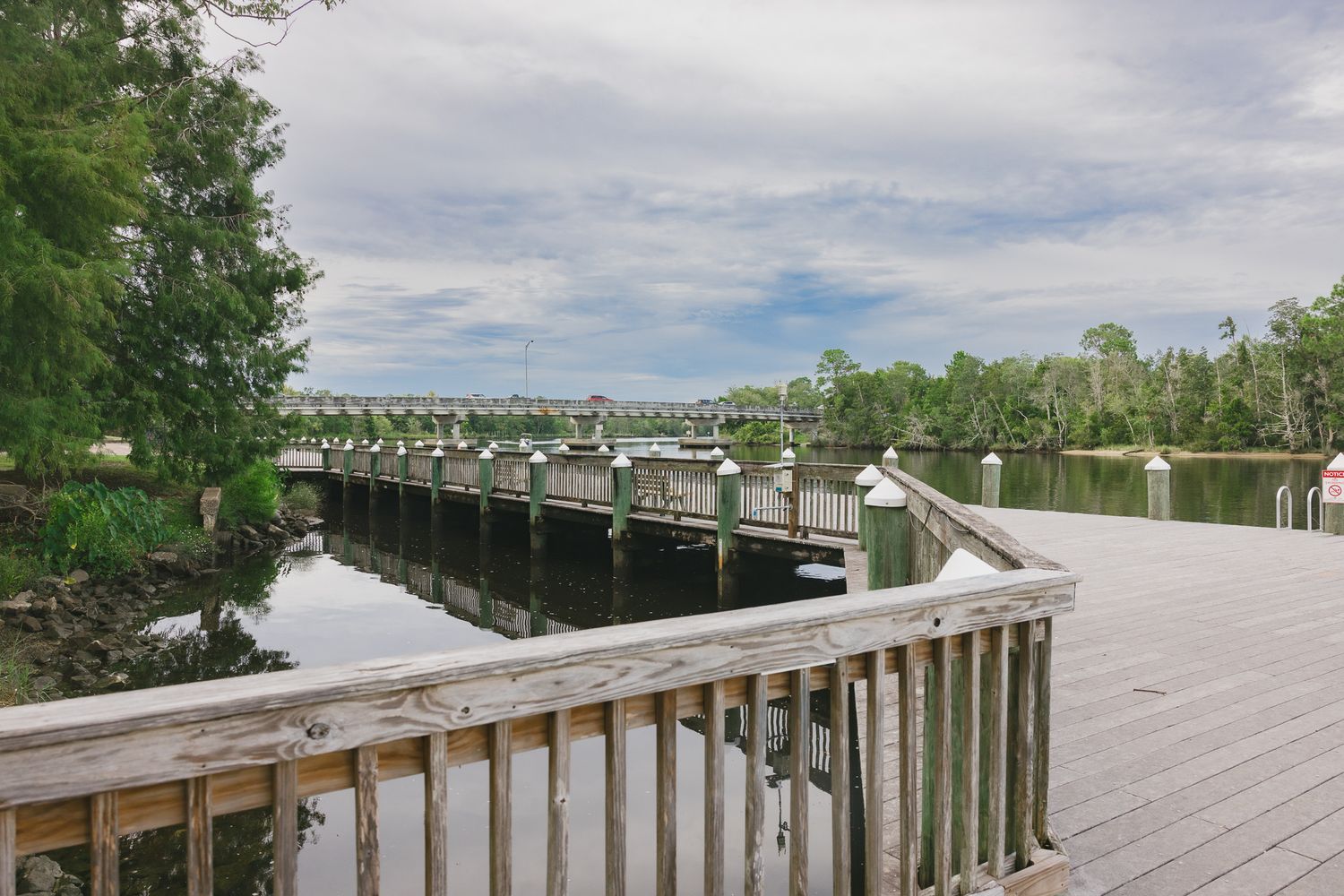 Wooden boardwalk and dock extending over calm water surrounded by lush green trees under a cloudy sky.