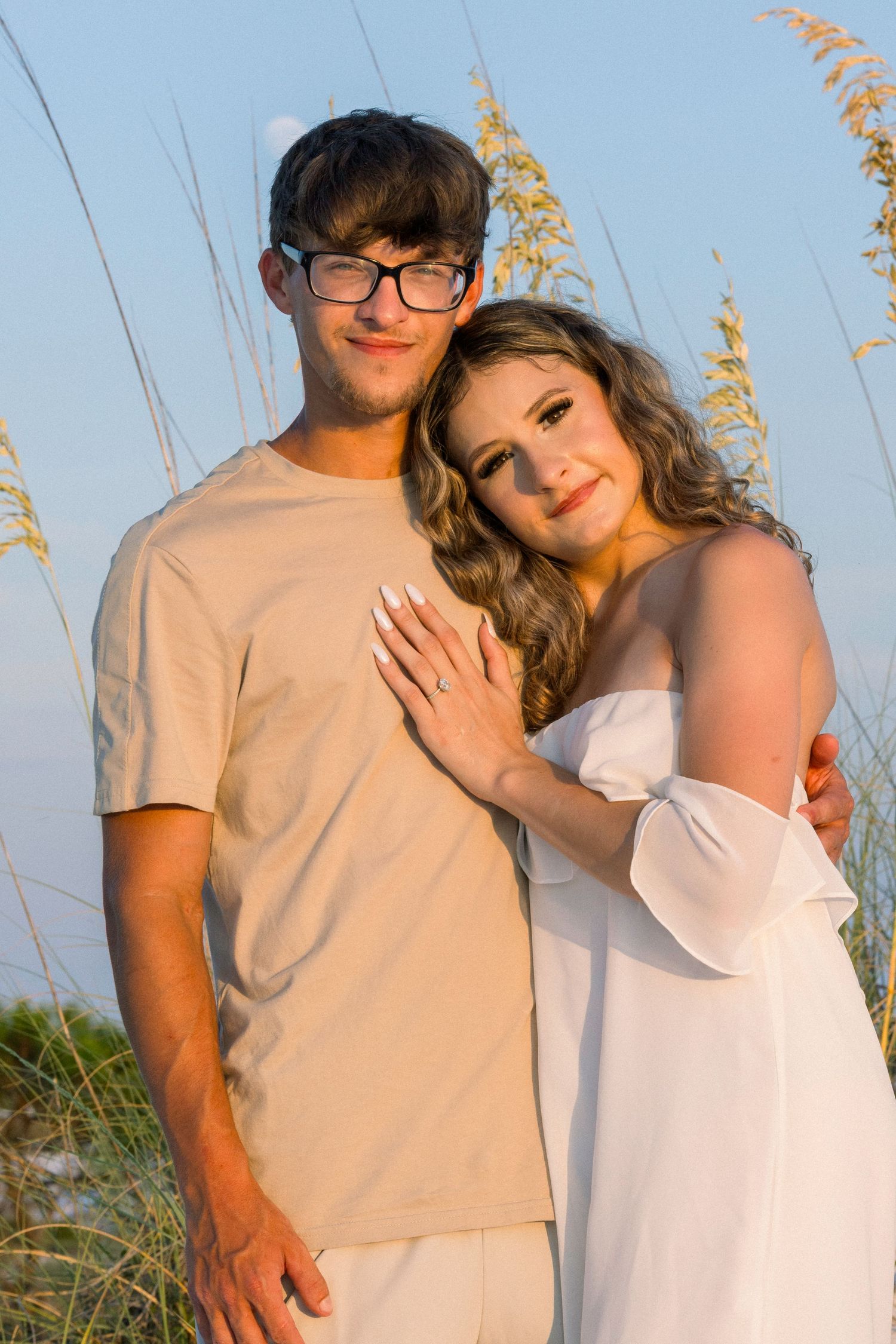 A romantic couple in light colored clothing poses together at sunset among beach grass with golden light.