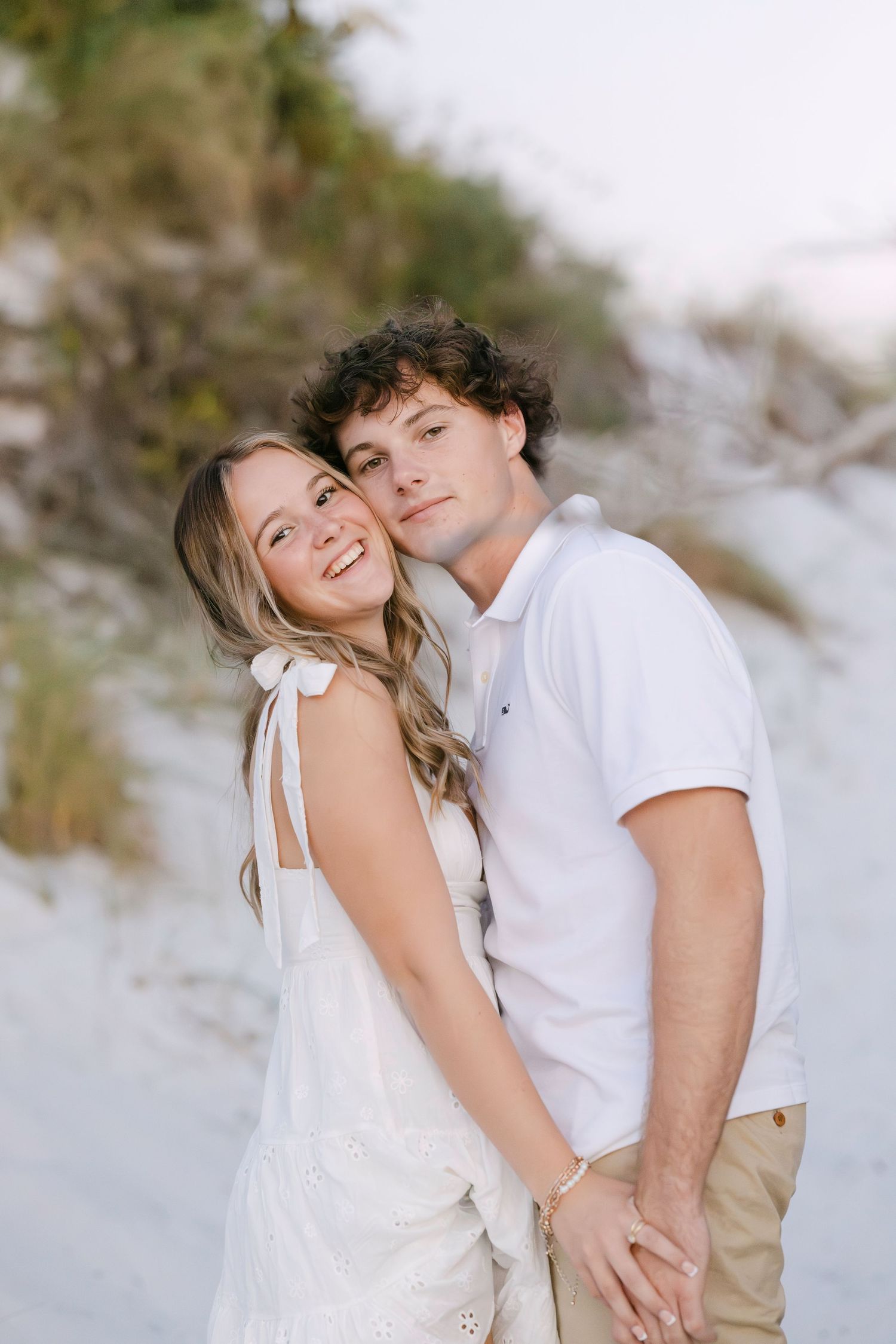 A romantic couple embraces on a sandy beach, wearing white outfits against a soft blurred background.