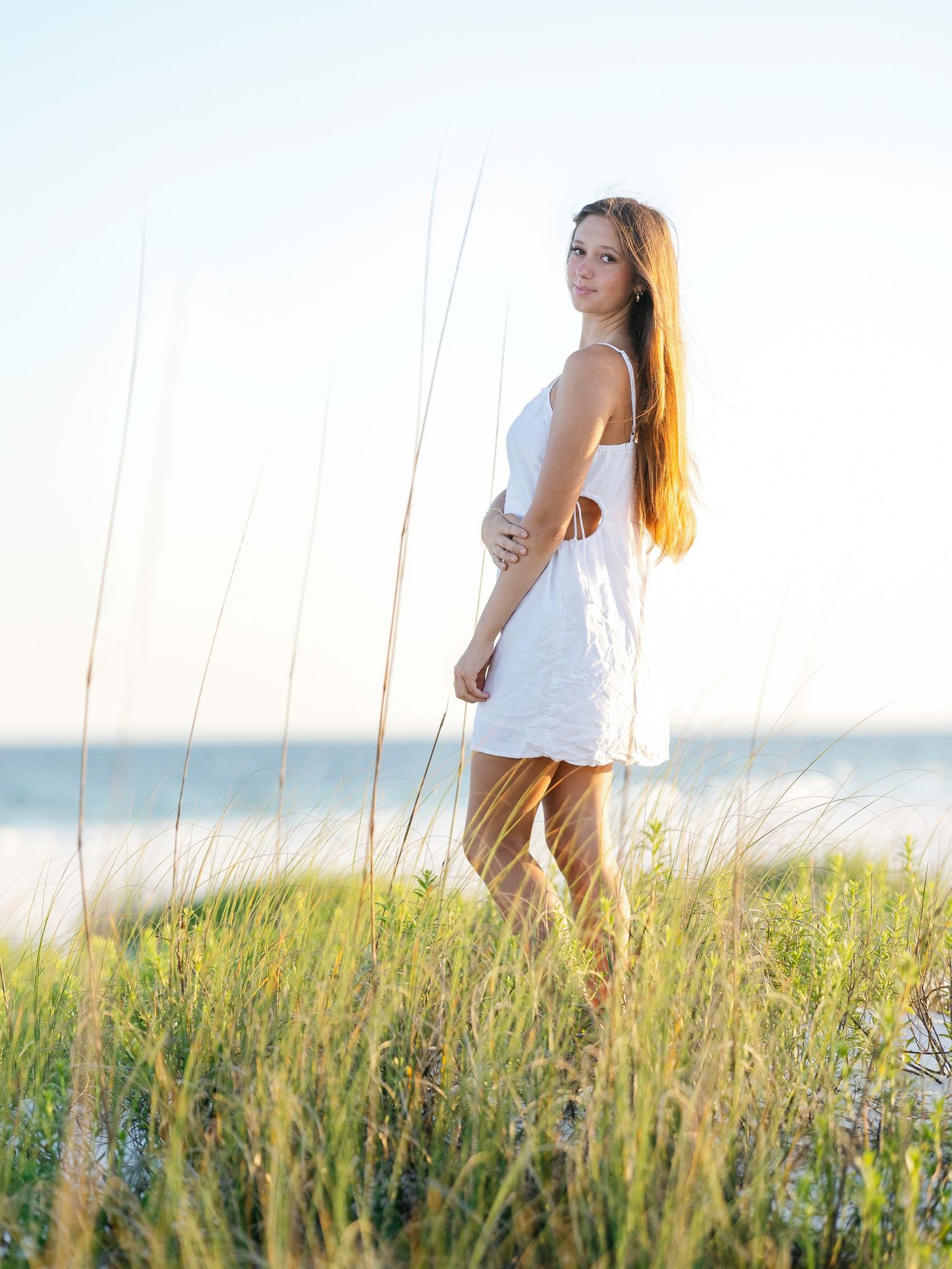 Person in white dress walking through tall beach grass during golden hour with ocean in background.