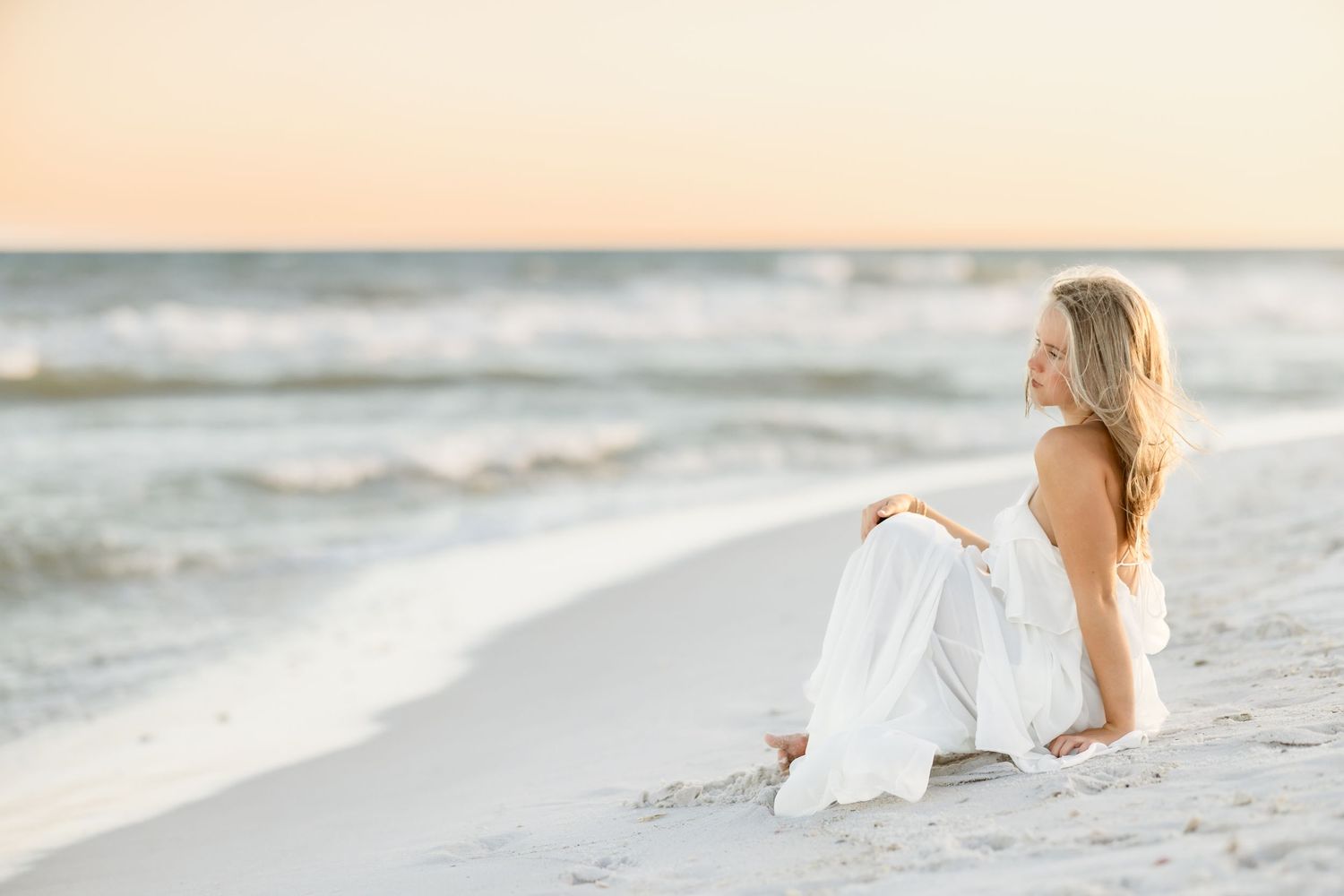 A blonde woman in a white dress sits peacefully on a sandy beach, gazing at the ocean during golden hour.