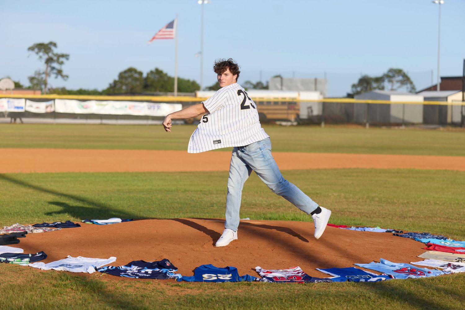 Young man in baseball jersey throwing a pitch on a decorated mound at an outdoor baseball field with American flag.
