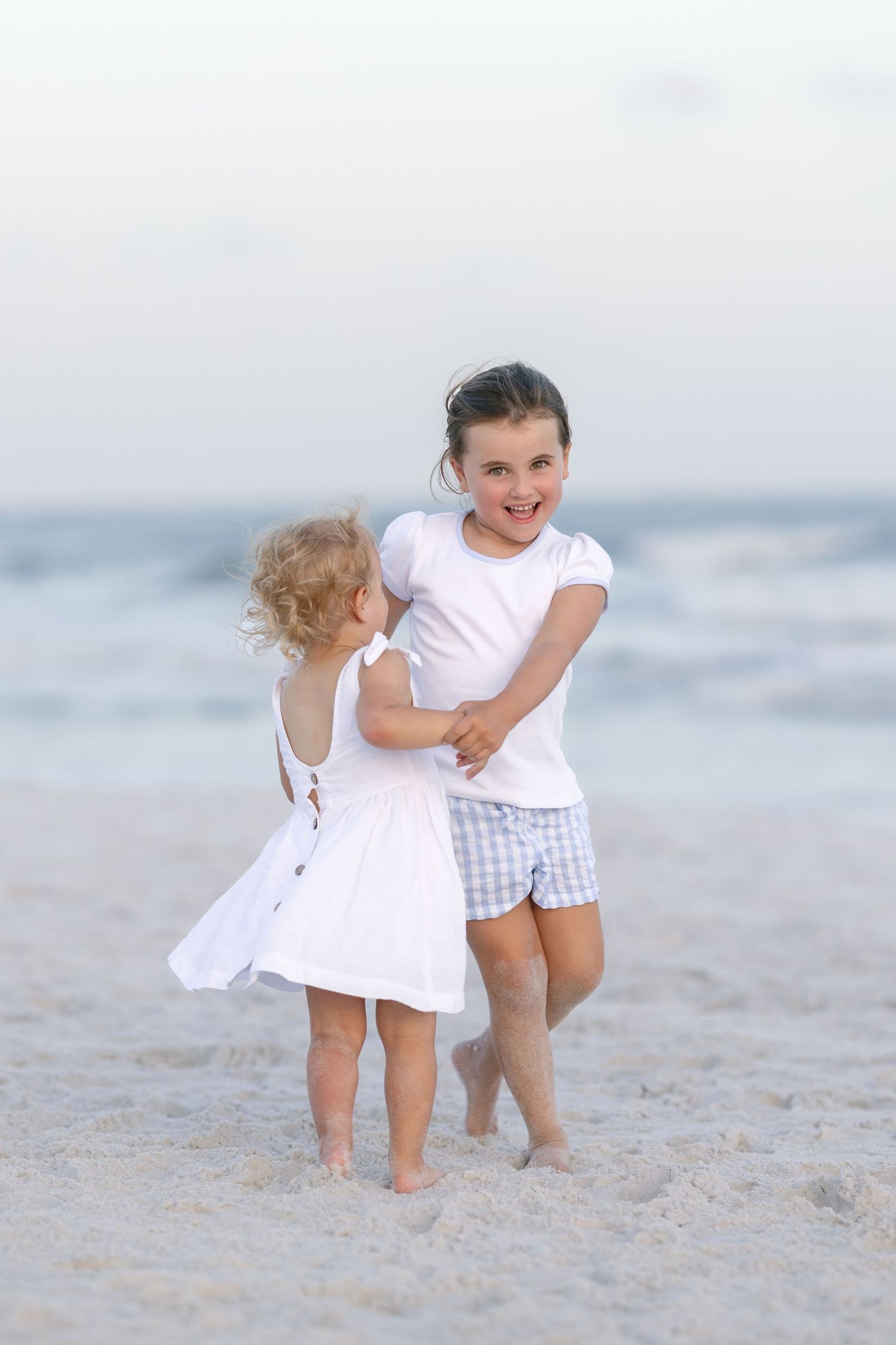 Two young girls playing joyfully on a sandy beach, one in a white dress, the other in a white top and plaid shorts.