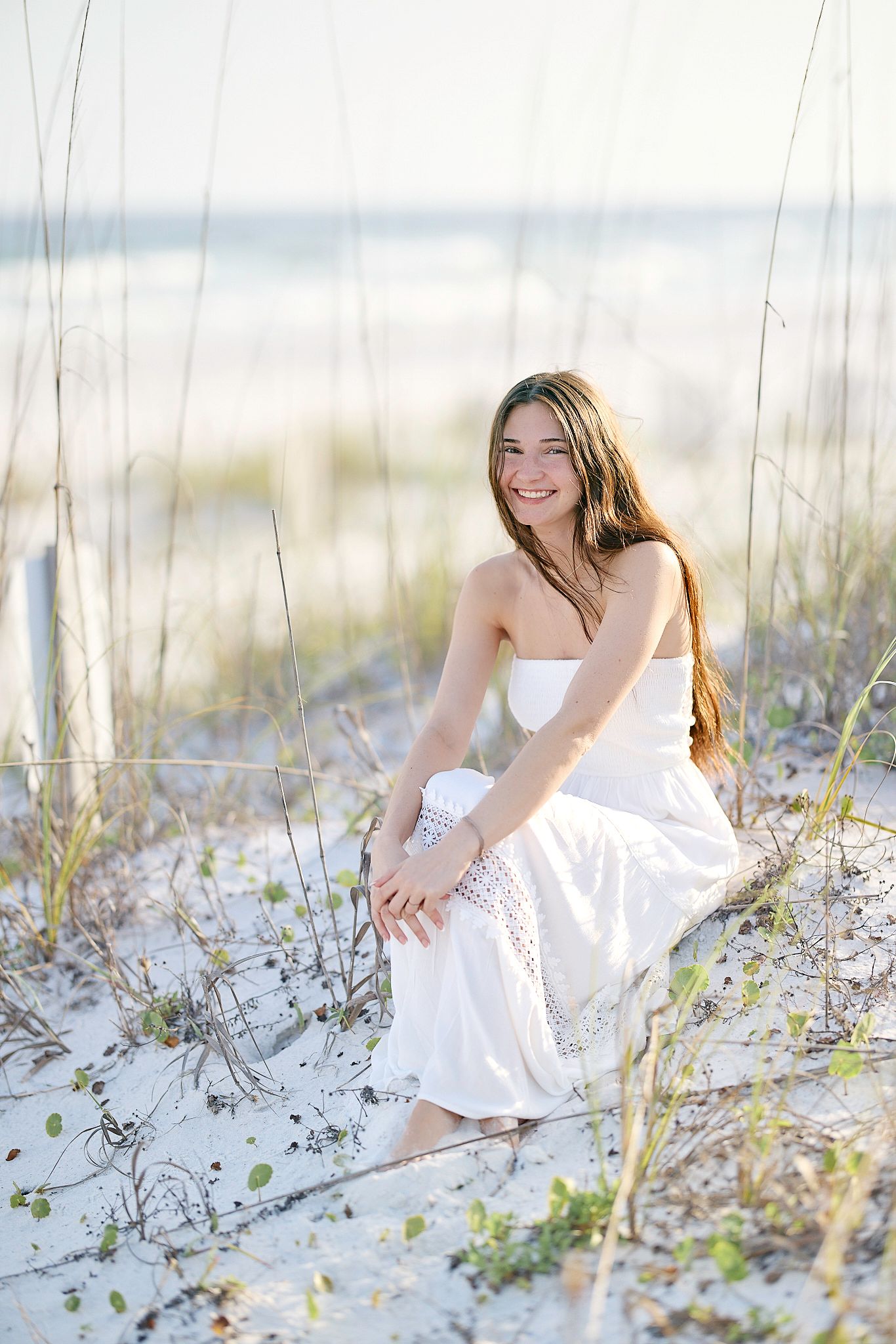 A casual beach photoshoot featuring someone in a white dress sitting among the dunes and sea oats.