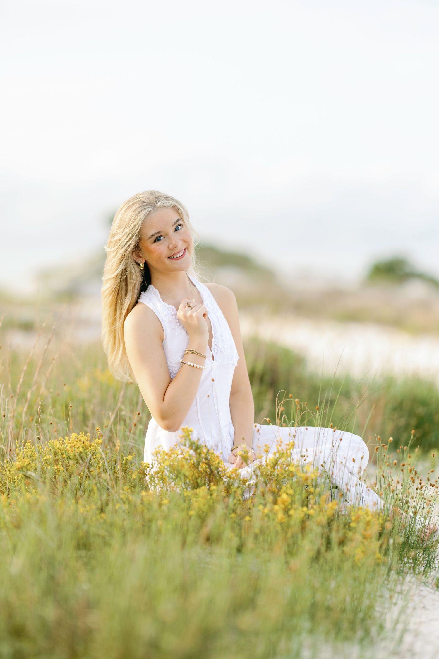 A woman in a white dress sits among wildflowers in a sunny meadow, creating a dreamy outdoor portrait.