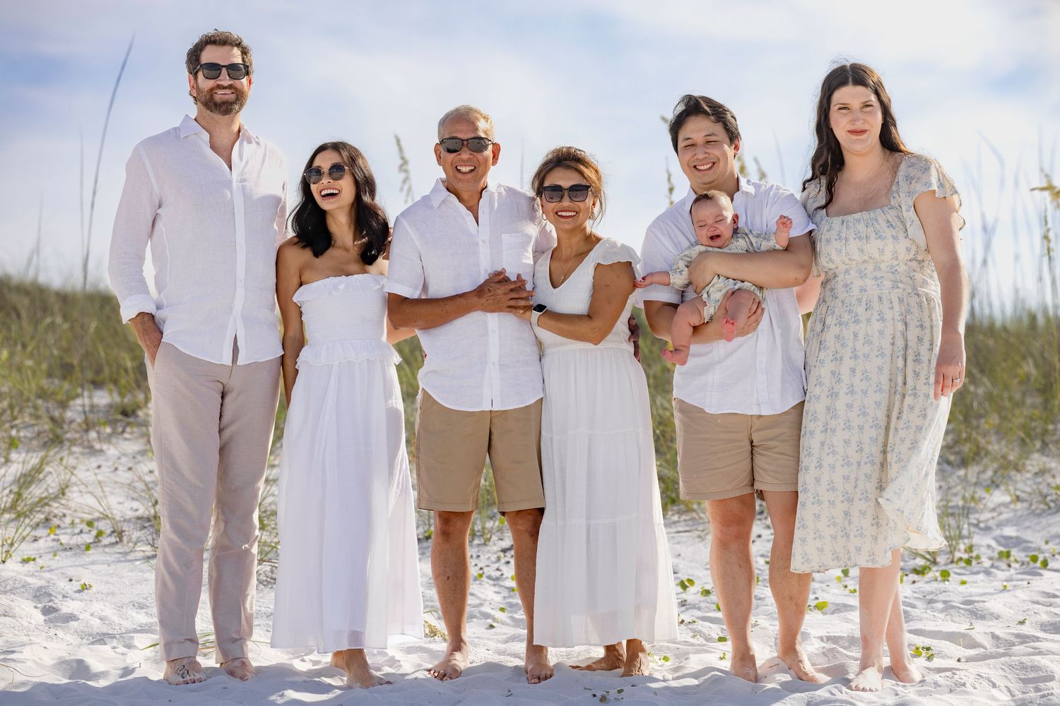 Family group photo on a sandy beach, smiling adults in white and tan outfits, holding a baby, with dunes behind.