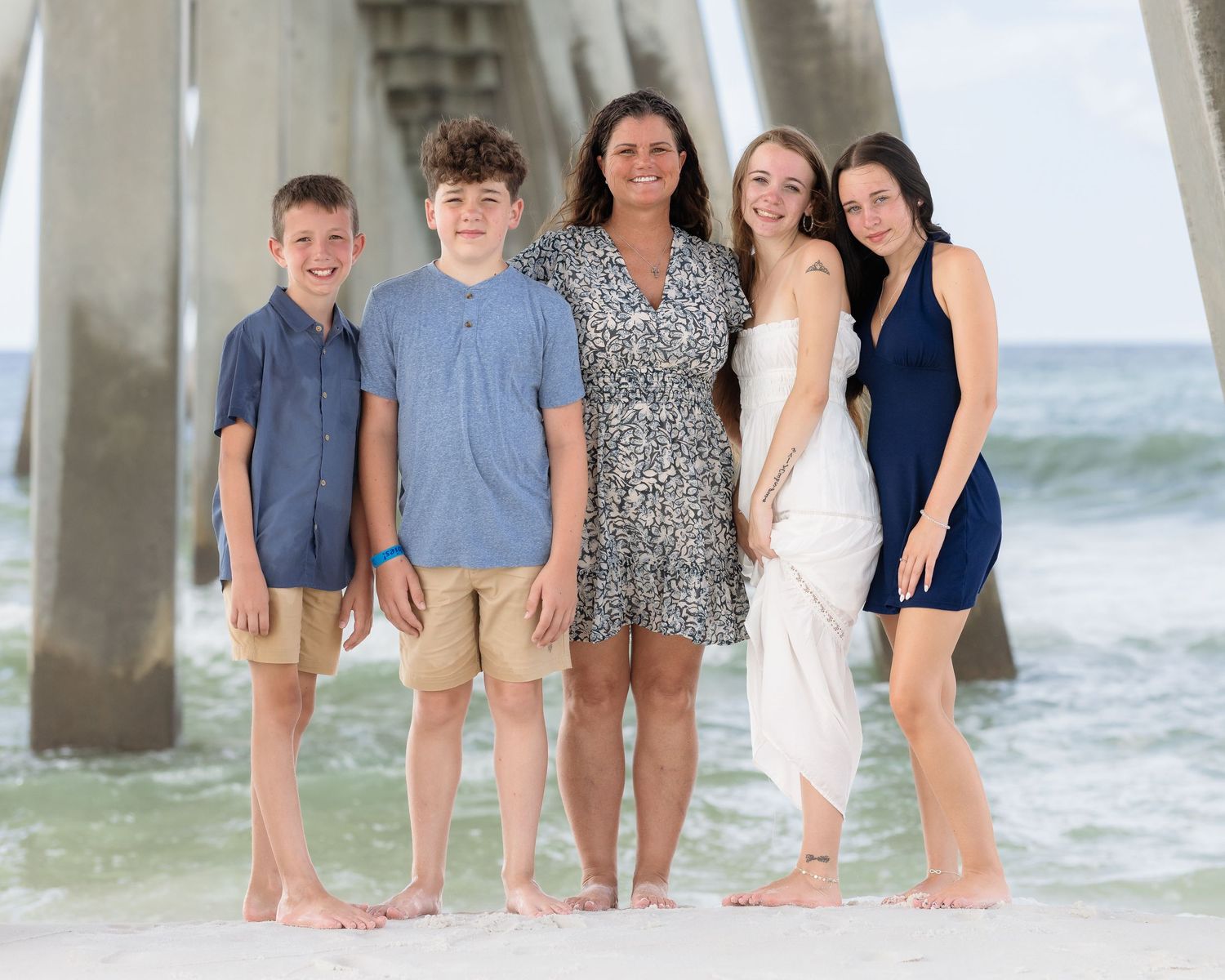 Family of five posing barefoot on sandy beach near pier, wearing casual summer outfits, smiling together.