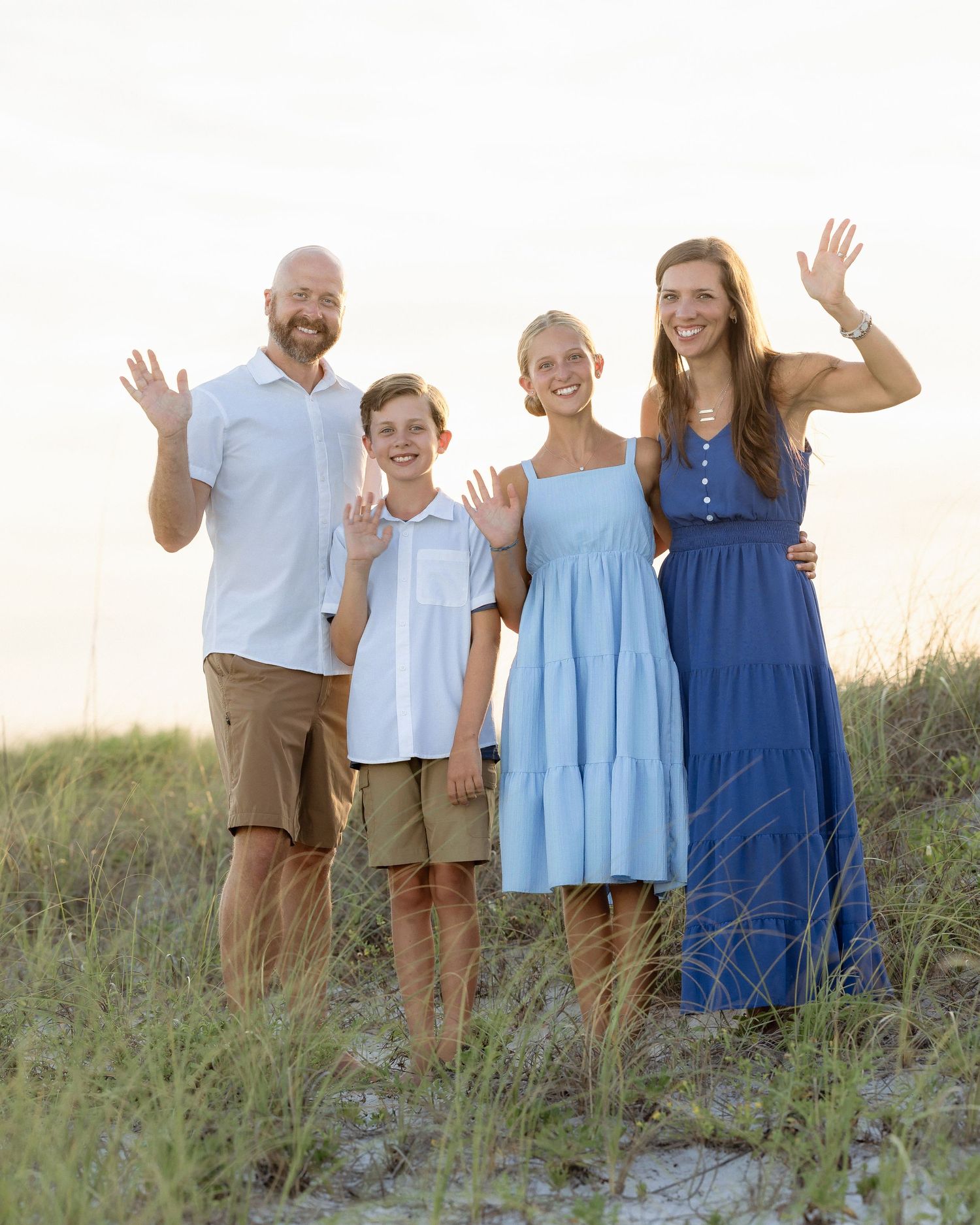 Family of four waving on a sandy beach at sunset, dressed in blue and khaki outfits, smiling joyfully.