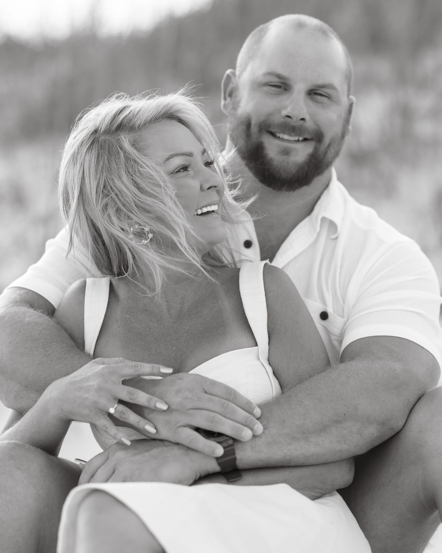 Black and white photo of a smiling couple embracing on a beach, woman in white dress, man in polo shirt.